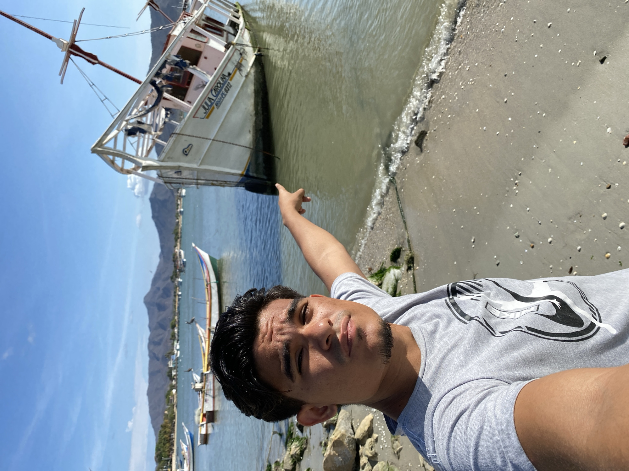 A bright, sunlit outdoor scene features a young adult man taking a selfie by a body of water, likely a bay, with mountains in the background. The man, with dark hair and a short beard, wears a light grey t-shirt with a distinctive black and white graphic design. He is positioned in the foreground, facing the camera with a neutral to serious expression, and is pointing his right arm towards a boat in the water.
To the man's left and slightly behind him is a large, white, wooden-hulled boat, partially submerged and resting close to the sandy and rocky shoreline. The boat has a mast and appears to be a traditional fishing or transport vessel. Clearly visible on its side are the words "JULIA CAROLINA" in yellow lettering, followed by "MISH.PELI.0212" in a smaller font.
The immediate foreground shows a mixture of sand and small rocks, with scattered seashells and gentle waves lapping at the shore. The water near the bank is a light, somewhat murky green, transitioning to a deeper blue further out. In the mid-ground, beyond the main boat, several other smaller boats are moored in the water, adding to the coastal ambiance. Majestic mountains dominate the distant background under a clear, bright blue sky with a few wispy clouds, suggesting good weather conditions during the daytime. The overall setting points to a vibrant coastal area in Juan Griego, Venezuela.