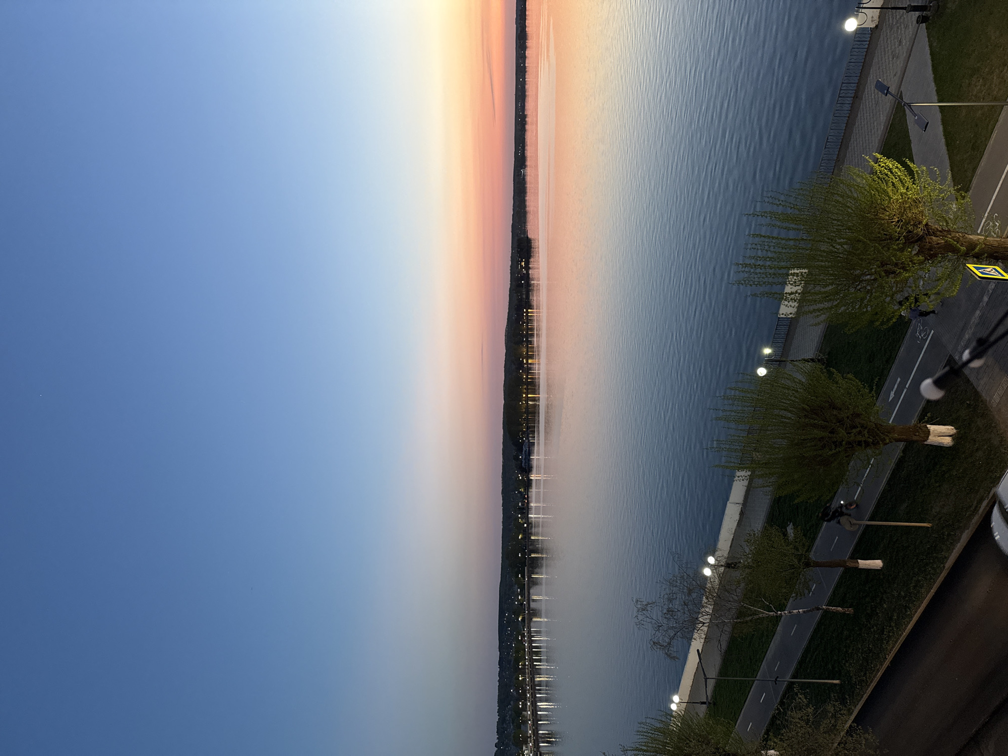 The image captures a scene along the Votkinsk reservoir in Votkinsk, Russia at dusk. The sky transitions from blue to orange near the horizon, reflecting in the calm water of the reservoir. A strip of land with lights runs along the reservoir edge, with reflections visible in the water. On the right side, a road lined with trees and streetlights parallels the reservoir. Two individuals appear to be walking along the road.