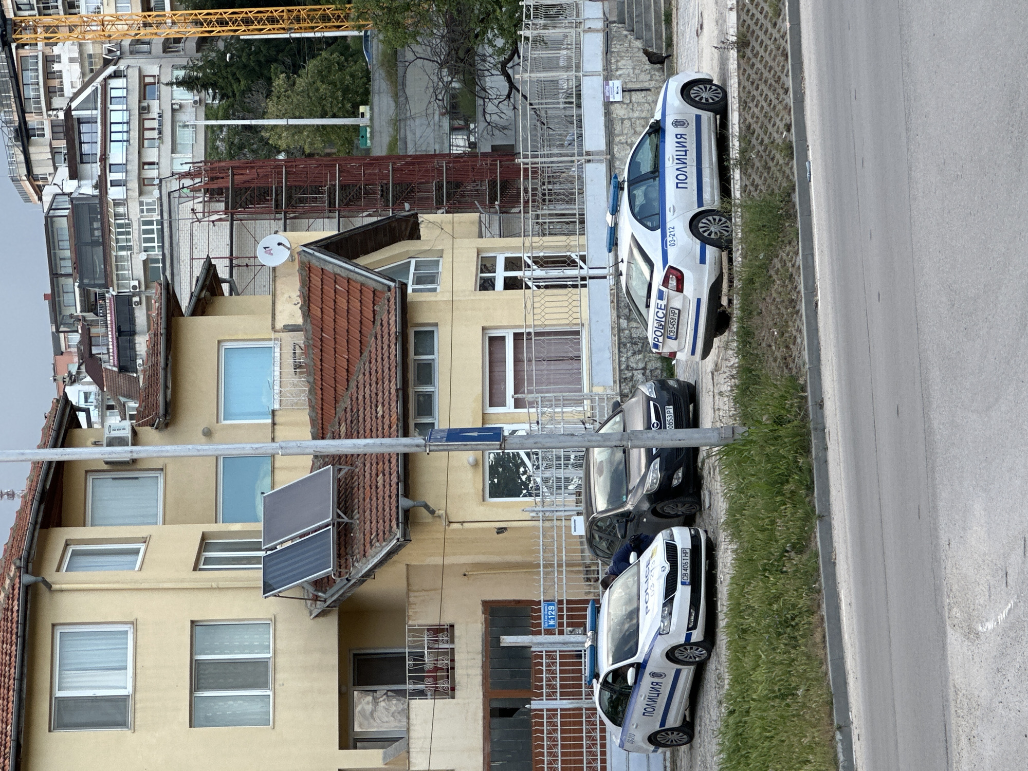 The image captures a street scene in Varna, Bulgaria, focusing on a row of buildings and parked vehicles. The buildings are primarily residential, exhibiting a mix of architectural styles, with yellow exteriors, tile roofs, and various window designs. A satellite dish is affixed to one of the buildings. A construction crane is visible in the background, suggesting ongoing construction in the area.
Two white and blue police cars are parked along the curb, facing opposite directions. Another black car is parked between the two police cars.
The image conveys a sense of everyday urban life. The scene appears to be taken during the daytime.