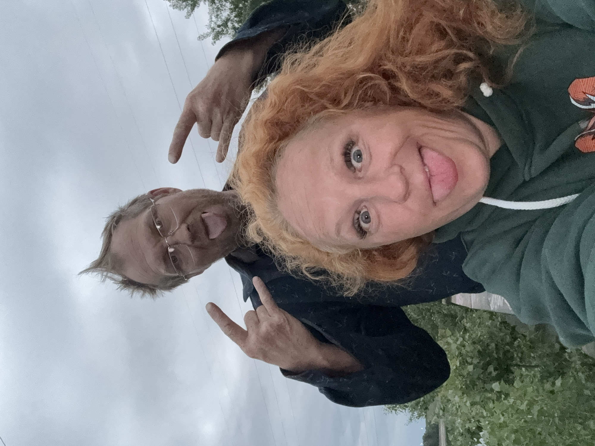 In a selfie, a woman with bright red, curly hair and wide, excited eyes sticks out her tongue. She is wearing a dark green hooded sweatshirt. Behind her, a man with spiky blond hair and glasses also sticks out his tongue. He is wearing a dark jacket and making a "rock on" hand gesture with both hands. They appear to be outdoors, with a cloudy sky and some greenery in the background. The setting suggests a casual, possibly rural or park-like environment, with a hint of power lines in the distance. The overall mood is playful and energetic.