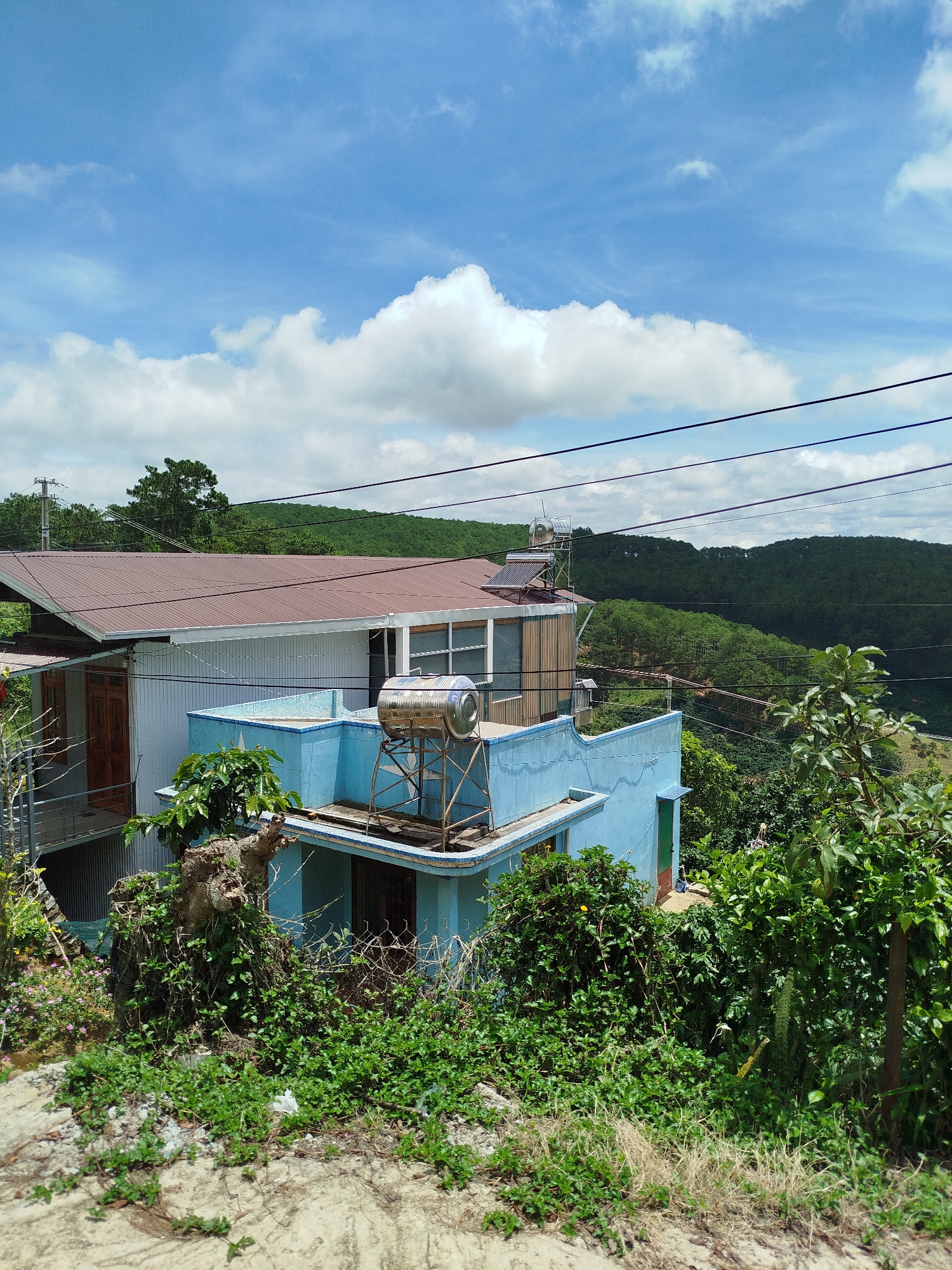 The image depicts a bright, sunny day with a clear blue sky and scattered white clouds. The scene is dominated by a two-story building, primarily painted in a pale blue. The lower level of the building is partially visible, with a green door and some overgrown vegetation. The upper level features a flat roof with a blue railing and a large stainless steel water tank mounted on a metal stand. To the left of the blue structure, a portion of another building is visible, characterized by a corrugated brown metal roof and white siding. Wooden doors and window frames are present on this structure.
The surrounding environment is lush and green, with dense foliage and trees covering the rolling hills in the background. The landscape suggests a rural or semi-rural setting, possibly on the outskirts of a city. Overhead power lines are strung across the scene, indicating the presence of infrastructure. The foreground consists of a dirt or gravel path with scattered dry grass and small green plants. The overall impression is one of a somewhat rustic residential area nestled within a verdant landscape. The image appears to be taken from an elevated position, looking down onto the buildings and the surrounding terrain. The specific location of Đa Thọ, Vietnam, is mentioned, suggesting this is a residential property within that area. No people are visible in the image.