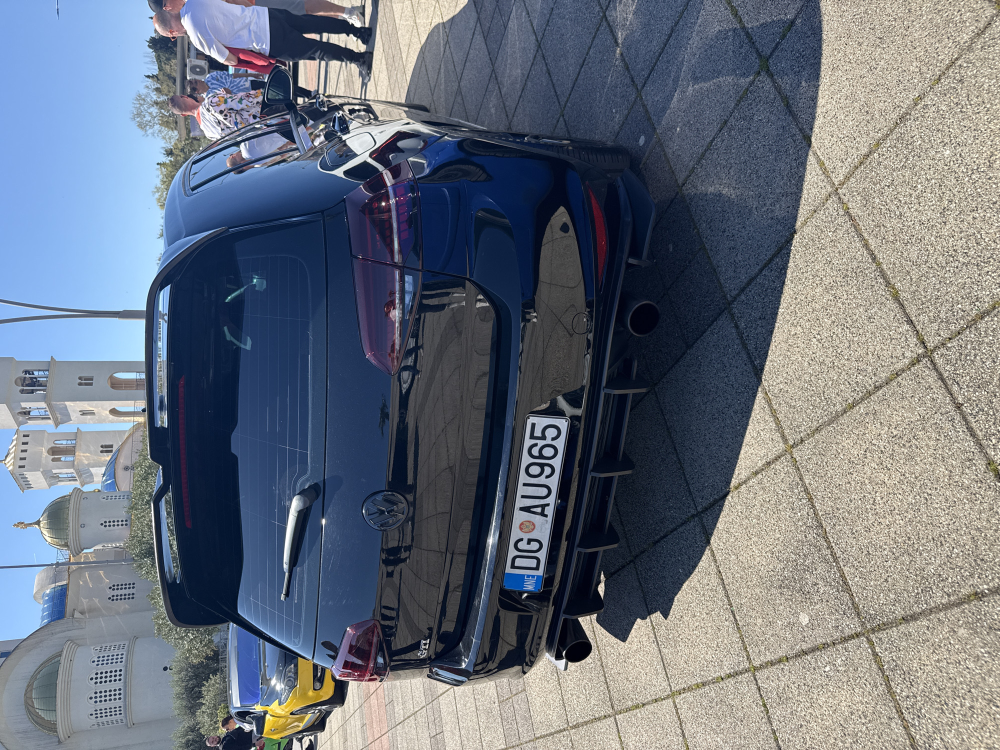 The image captures the rear of a black Volkswagen GTI with the license plate "DG AU965." The car is parked on a paved surface. In the background, there is a white and blue building with a dome. There are multiple people visible, mostly in the upper part of the frame.