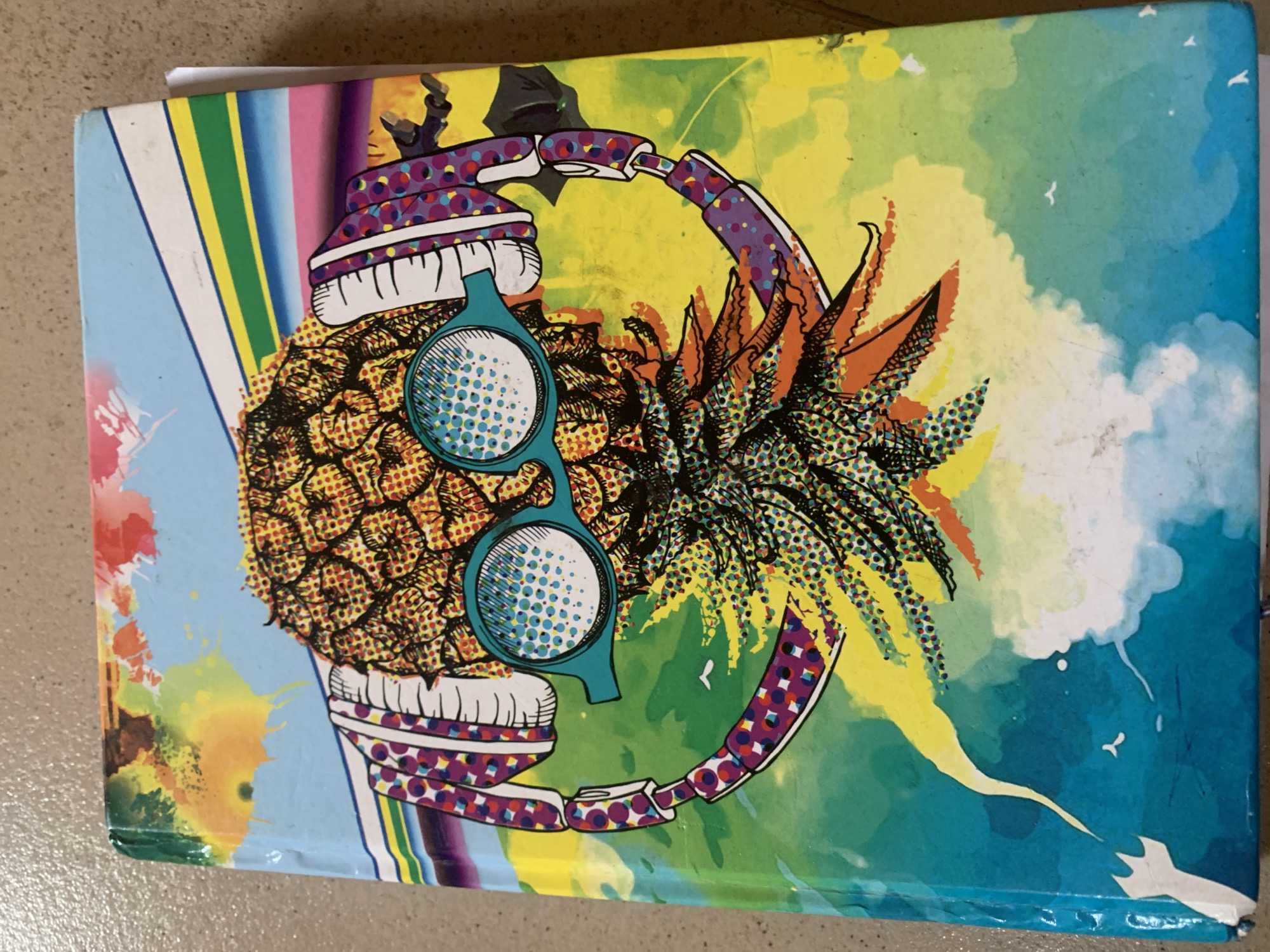 The image shows the cover of a notebook with a vibrant and abstract design. The central subject is a stylized pineapple wearing headphones and oversized sunglasses. The background features a colorful splash of paint and abstract shapes in shades of blue, green, yellow, and orange, with diagonal stripes in various colors at the top. There are also small silhouetted figures of what appear to be people in the upper right corner of the notebook cover, one appearing to be leaping. The notebook is lying on a textured surface that appears to be a countertop or table. The image is taken from a top-down perspective, slightly angled. There are no specific landmarks or details that indicate a city or location within a city.