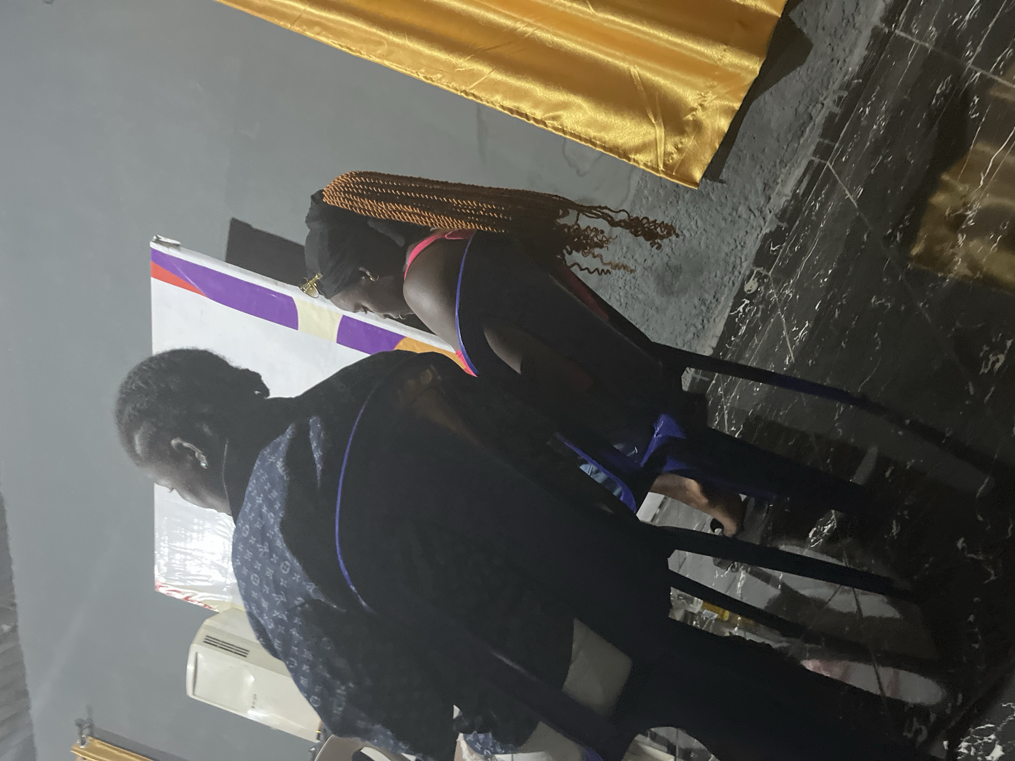 The image shows two individuals, likely women, in an indoor setting. The person in the foreground is wearing a dark blue patterned garment with blue trim, and appears to be seated. The person in the background has long, braided hair and is wearing a dark top with pink trim, and is also seated. They are positioned near a white and purple banner or display. The room has dark, tiled flooring with white veining, and a gray wall. A golden fabric hangs from the top of the image, possibly a curtain or drape. An air conditioning unit is visible below the banner. The lighting suggests it is evening or indoors with artificial light. The image is oriented vertically, with the top of the scene appearing on the left side.