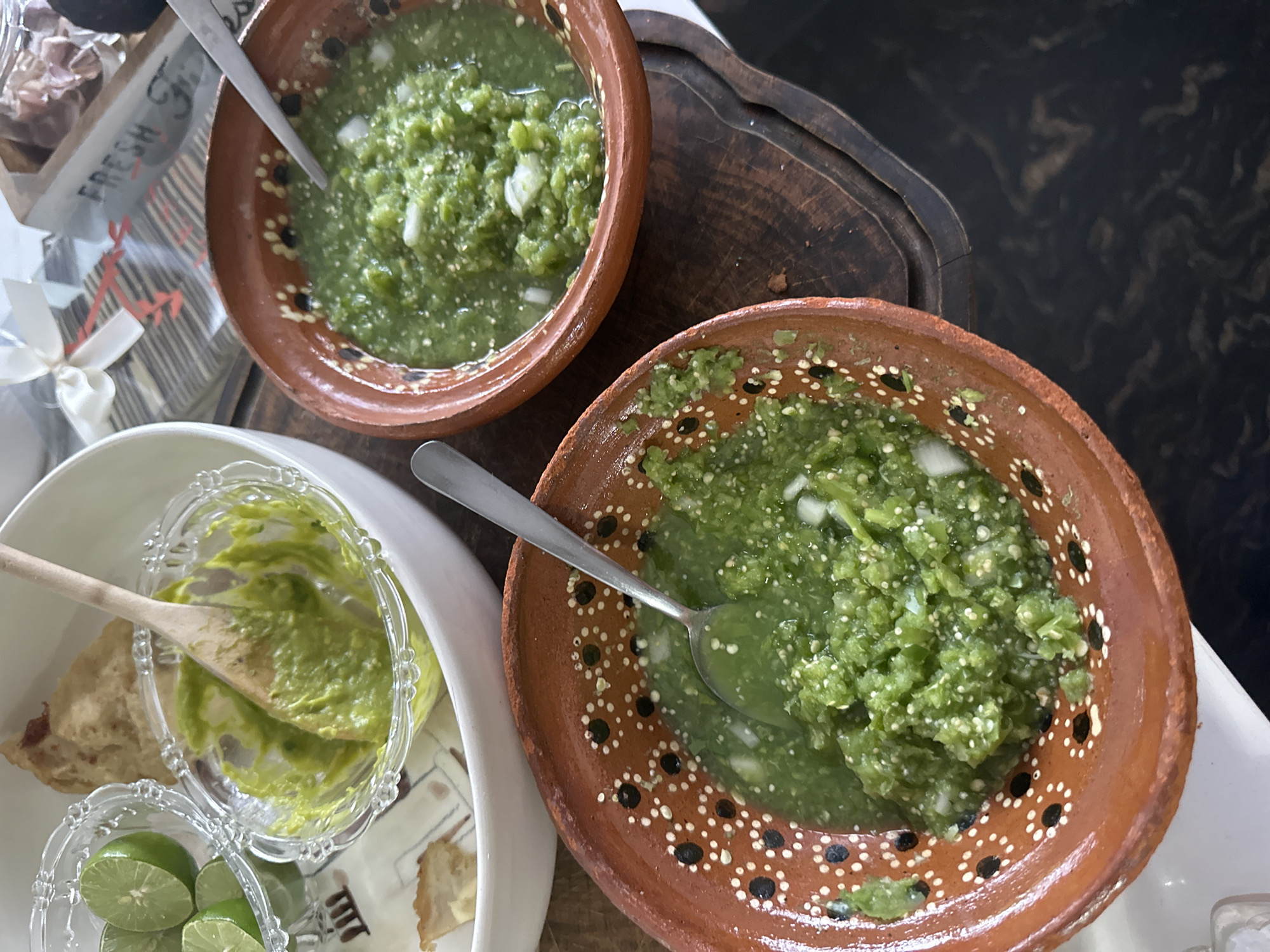The image shows a table setting with Mexican food, likely in Naucalpan de Juárez, Mexico. There are two rustic brown clay bowls filled with green salsa, each containing a metal spoon. Nearby is a white plate holding a glass dish of guacamole and a small glass dish filled with lime wedges. Fried tortillas are visible beneath the dishes. A wooden serving tray sits behind the salsa bowls. A container marked "FRESH" is also in view. The table surface appears to be patterned or mottled.