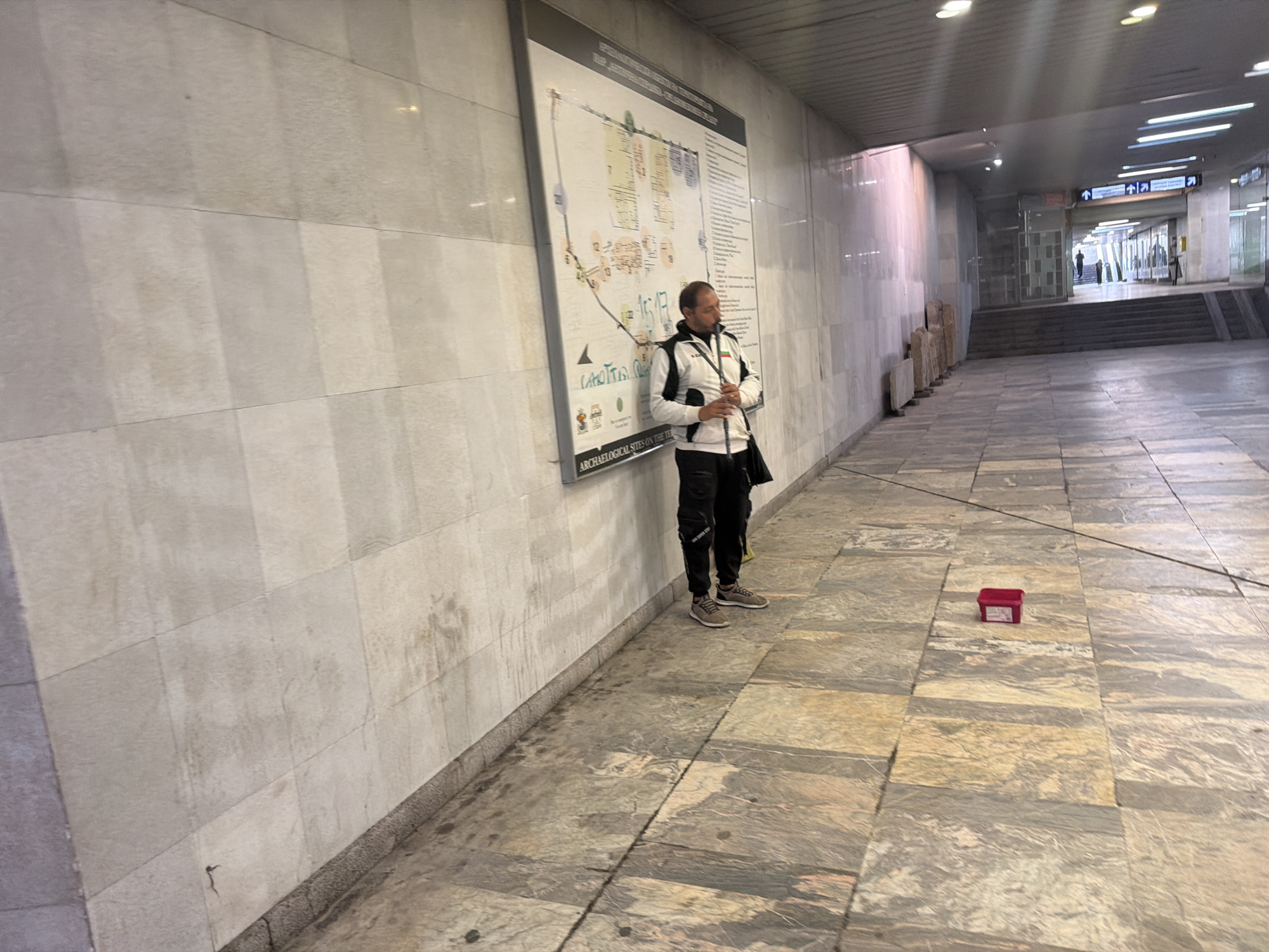 The image shows an indoor public space in Sofia, Bulgaria, likely a subway station or underground pedestrian area. A man stands near a large wall-mounted map labeled "ARCHAEOLOGICAL SITES ON THE TERRITORY OF SERDICA." He wears a white and black jacket, black pants, and sneakers, holding something in his hands. A red box sits on the patterned stone floor nearby. In the background, a passageway leads to stairs and a distant exit with overhead directional signs. The walls are light-colored stone. The lighting is subdued, creating a somewhat sterile atmosphere.