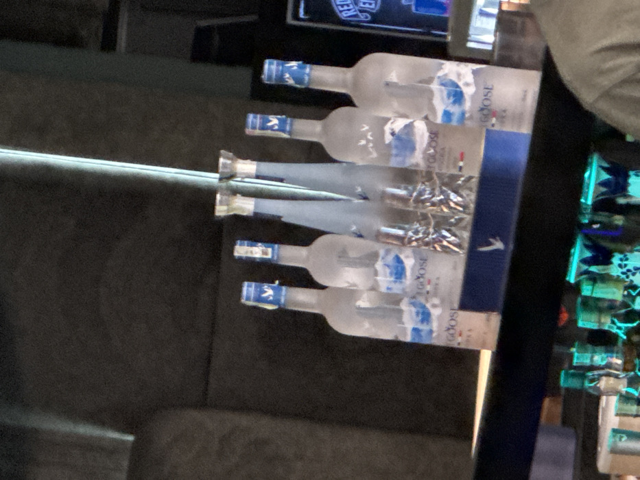 The image displays an indoor bar setting, focused on a display of six Grey Goose vodka bottles. The bottles are arranged on a dark, reflective surface, likely a bar shelf or counter. Five of the bottles are standard Grey Goose vodka bottles, identifiable by their frosted appearance, blue labels featuring a snowy mountain scene, and blue caps with a white goose logo. Two slender, clear bottles, potentially part of a special presentation or a different product line, are positioned in the center. Between these two central bottles, two clear, textured glasses are visible. A blue display stand with a white goose silhouette logo supports some of the bottles in the foreground. The background is dark and out of focus, featuring a vertical illuminated strip, possibly an LED light, running from the upper left to the lower right. Below the main display, a section of a lower bar shelf is visible, containing several bottles with green and blue accents, illuminated by a distinct neon-green light. A partial view of a person's shoulder and upper arm, wearing a light-colored garment, is visible in the bottom right corner of the frame. The specific location within Ruse, Bulgaria, is not discernible from the visual information presented in the image.