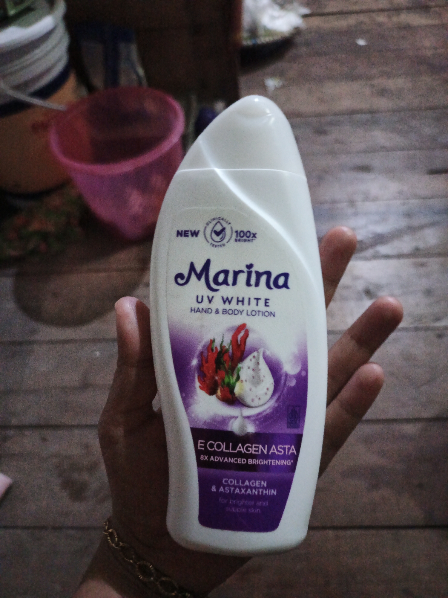 A close-up shot captures a person's hand holding a white plastic bottle of Marina UV White Hand & Body Lotion. The bottle is partially obscured by the fingers and thumb of the right hand, which exhibits a gold-colored bracelet around the wrist. The lotion bottle features a purple and white label with text and graphics. In the blurred background, a pink plastic cup and other indistinct objects are visible. The setting appears to be indoors, with a wooden surface in the background. The image does not provide any specific information about a location in Sinar Bulan, Indonesia.