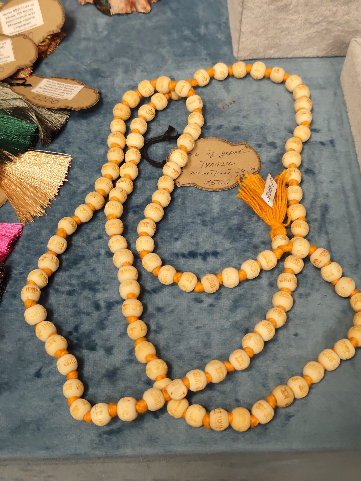 A close-up, overhead shot shows a mala, a string of prayer beads, made of light-colored wooden beads strung on an orange cord. Each bead is carved with golden Hindi script. The mala is laid out on a textured, dark blue fabric surface. To the left of the mala are several small, light brown cardboard tags, some with handwritten text in Hindi. One tag is shaped like a cloud and has the text "Бусы из дерева Туласи мантры Сита 1500". Other tags have more text in Hindi and some multi-colored tassels are visible below them, including gold, green, and pink. In the upper right corner, a corner of a light grey textured box is visible. This image was taken in Sochi, Russia.