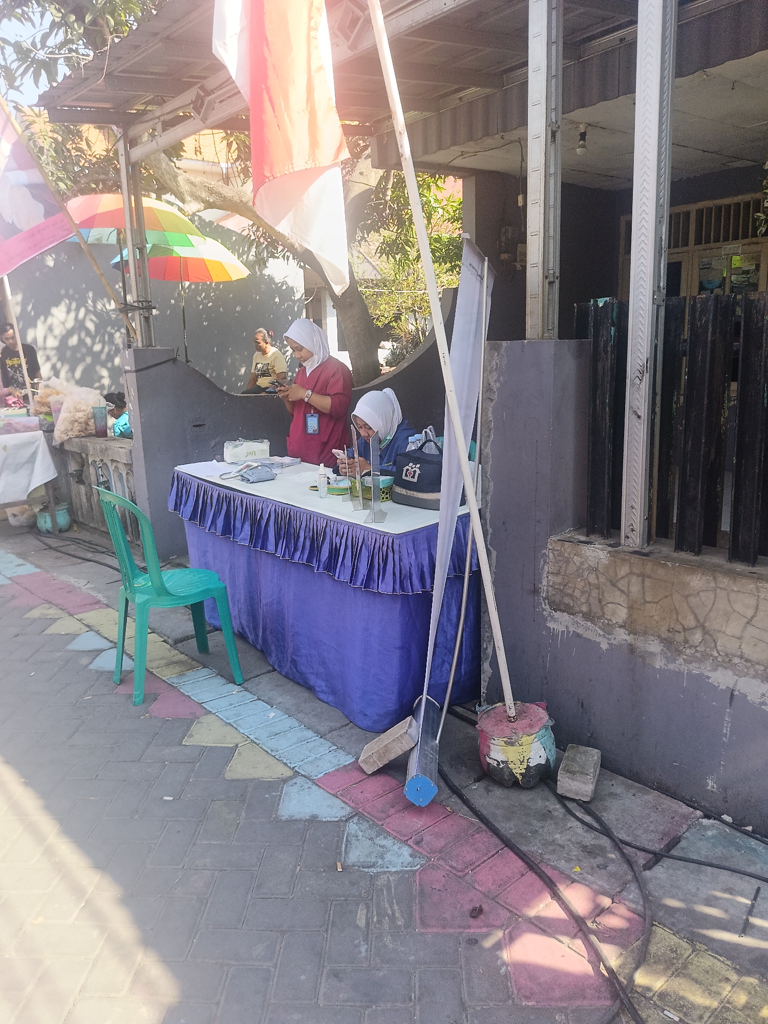 In Gedangan, Indonesia, an outdoor scene unfolds under a bright sky. The foreground features a paved walkway with colorful rectangular tiles. To the left, a green plastic chair sits empty. Dominating the center is a long table draped with a purple cloth, adorned with a ruffled edge. On the table, various items are present, including a box of tissues, a blue tissue holder, hand sanitizer, and what appears to be a menu or list. Two women, both wearing hijabs, are attending the table. One, in a maroon top, is looking down at her phone, while the other, in a blue top, is also engaged with a device. Behind the table, a large gray wall serves as a backdrop, with a black metal fence to its right. Colorful flags, including Indonesian red and white, are visible in the upper portion of the image, along with some tropical foliage. Sunlight casts shadows across the scene. To the right of the table, a broom with a blue tip leans against a colorful, painted object that appears to be a repurposed bottle or container, with a brick nearby. Further to the left, another table is set up with bags of snacks.
