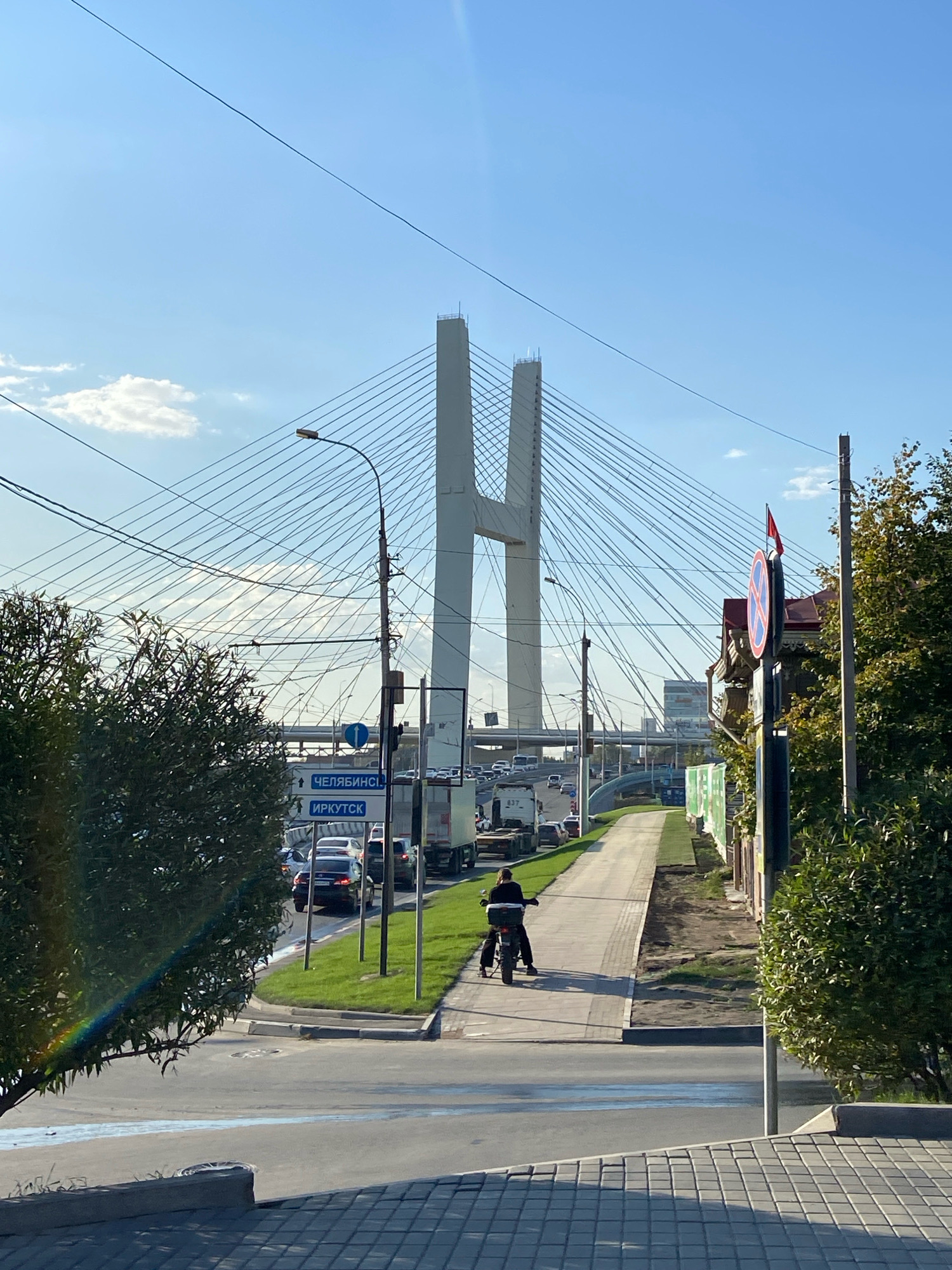 The image depicts a sunlit scene in Novosibirsk, Russia, featuring a prominent cable-stayed bridge. In the foreground, a paved area transitions to a sidewalk where a person is riding a motorcycle. The sidewalk is bordered by grass and a road with several cars and trucks, indicating traffic. Two blue street signs point in opposite directions, one indicating "CHElyabinsk" and the other "IRKUTSK". A traffic sign depicting a red circle with a diagonal red bar over a blue background is visible on the right side of the frame. Trees with green foliage frame the scene, and power lines are strung across the sky. The sky is blue with some wispy clouds. The bridge, a large structure with two towers connected by cables, dominates the background.