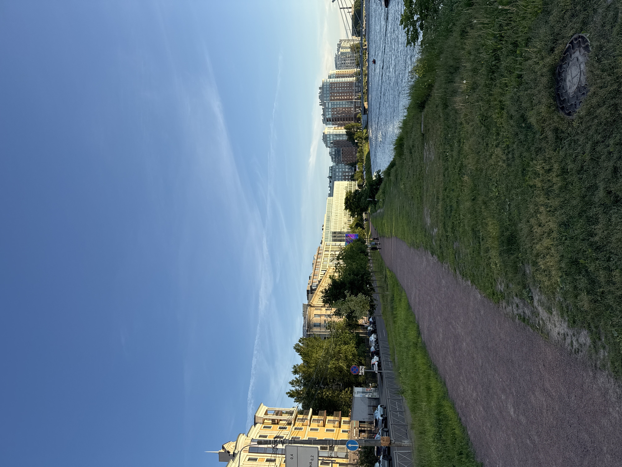 This is a photograph of a waterfront path in Saint Petersburg, Russia, likely taken in the evening as the sun casts a warm glow on the buildings. The path, made of dark red gravel, runs along the grassy bank of a wide river, its surface rippling under the clear blue sky.
On the left side of the path, a tree-lined street runs parallel to the river. A row of modern apartment buildings with varying architectural styles, from classical to contemporary, line the street. Some of these buildings are painted in warm yellow and beige tones, while others feature red brick or glass facades. Cars are parked along the street, and a few people are visible walking on the sidewalk.
Further down the river, a bridge spans the water, and more buildings can be seen on the opposite bank. The overall impression is one of a well-maintained urban environment that blends modern living with natural scenery.