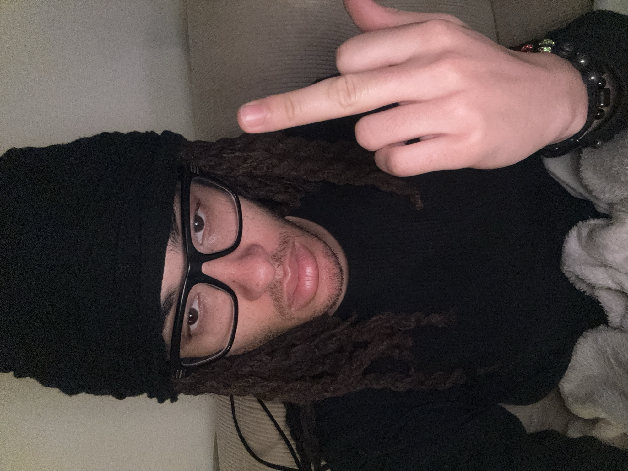 A man with dreadlocks and glasses, wearing a black beanie and black shirt, lies on what appears to be a beige couch. He is giving the middle finger. He has a beaded bracelet on his right wrist. The scene does not provide any information to suggest that it is in Paris, France.