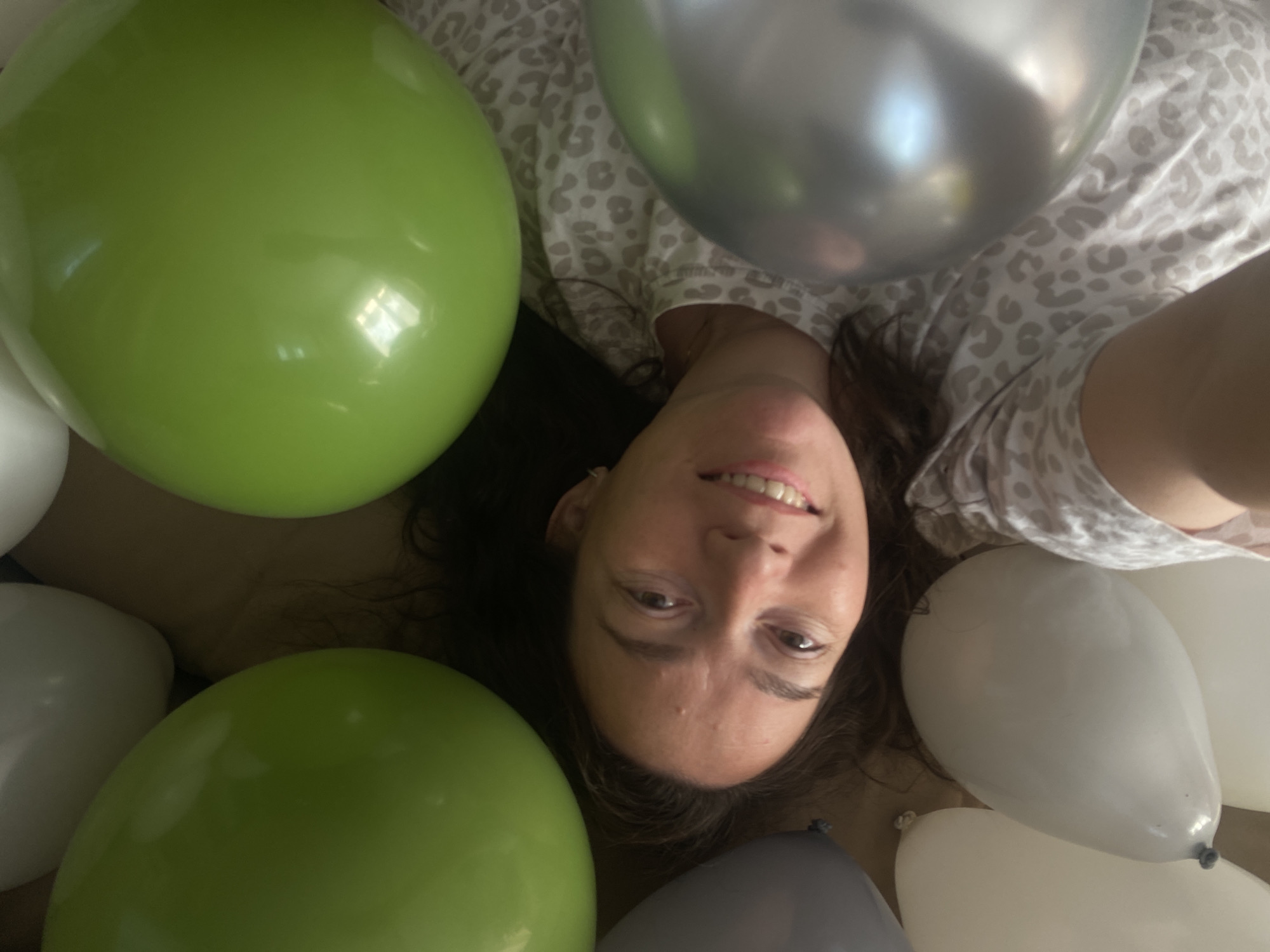 A woman lies on her back, surrounded by several balloons in shades of green, gray, and white. She is wearing a patterned, light-colored top. The location is unknown, and there is no indication that it is in Ольховка, Russia.