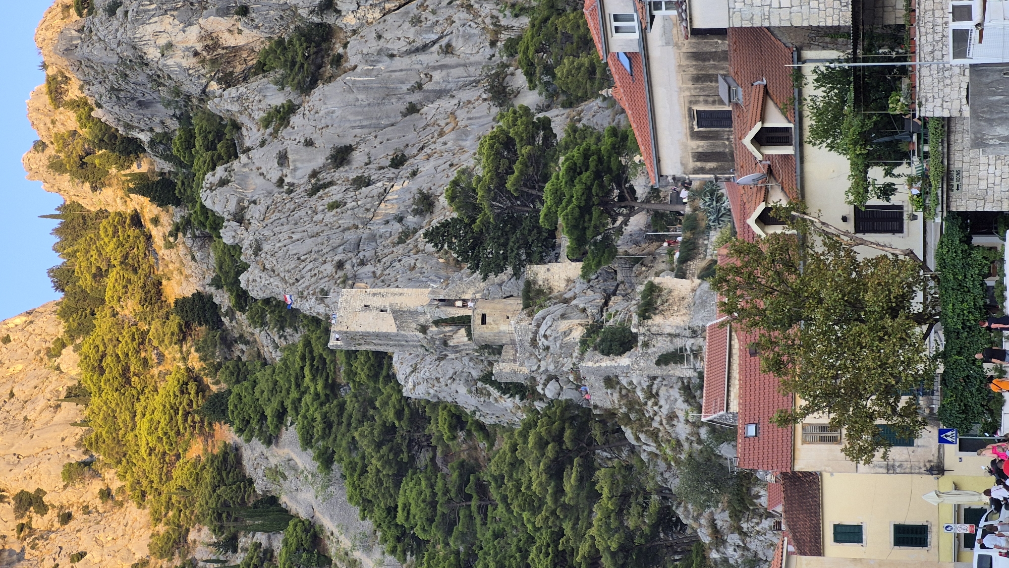 The image depicts a medieval fortress perched precariously on a steep, rocky mountainside, overlooking a cluster of red-roofed buildings in the foreground. The fortress, constructed from weathered stone, features a prominent tall tower and several lower structures, some of which are partially in ruins. Lush green trees and dense vegetation cling to the rocky slopes, contrasting with the stark gray of the mountain. The sky above is a clear, pale blue. The buildings in the foreground are of a traditional Mediterranean style, with cream-colored walls and red-tiled roofs. Some of these buildings have visible windows and balconies, and a few people can be seen on the street level. The overall impression is one of historical grandeur set against a dramatic natural landscape.