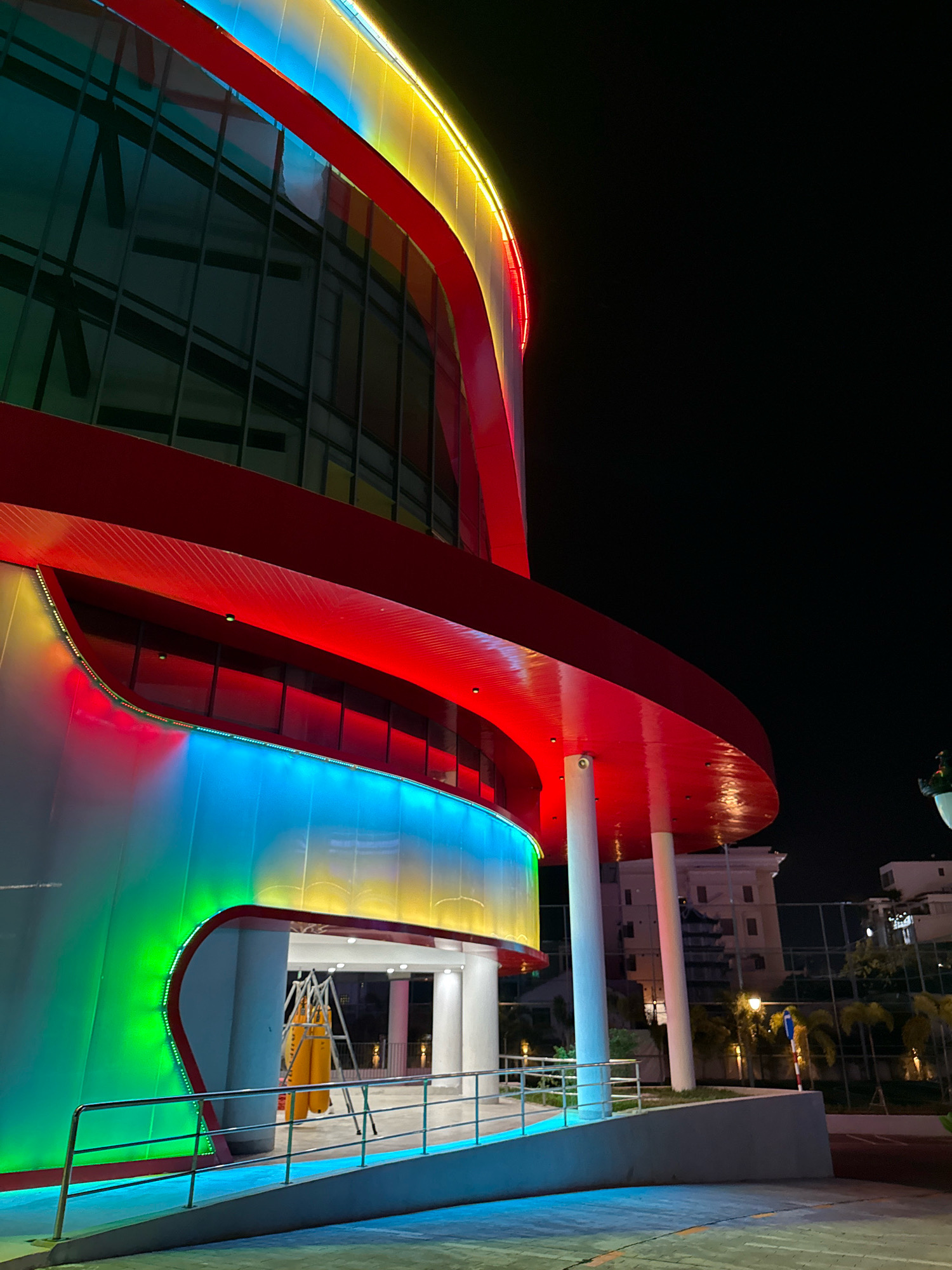 The image depicts a modern architectural building with vibrant, colorful LED lighting at night, located in Nha Trang, Vietnam. The building features a curved facade with large glass windows and is illuminated by bands of red, blue, green, and yellow lights that wrap around its exterior. The overall aesthetic is futuristic and striking. In the foreground, a paved walkway with a metal railing leads towards an entrance. Inside the visible entrance, there are several yellow punching bags suspended from a metal frame. Beyond the building, the night sky is dark, with a few distant lights and trees visible. The architectural style suggests a public facility or entertainment venue.