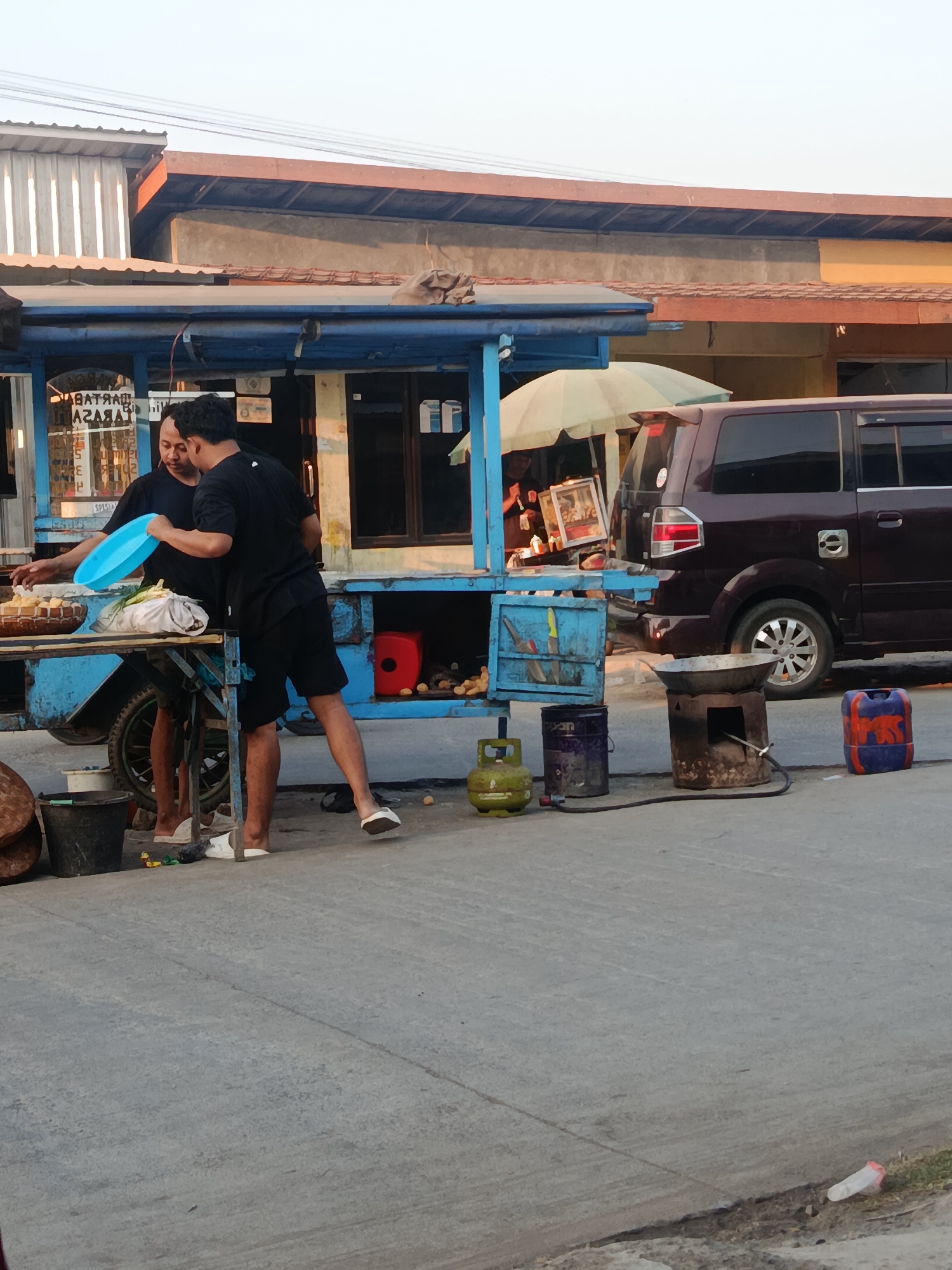 The image depicts a street scene in Kedungmutih, Indonesia, with a food stall as the central focus. Two men are present, appearing to be interacting with food preparation items. One man, wearing a black t-shirt and shorts, holds a blue plastic bowl. The other man, also in black attire, stands beside him, looking towards the food. The food stall itself is constructed from blue metal and appears to be a mobile cart. Various items are visible around the stall, including buckets, a gas cylinder, a metal stove with a wok, and a maroon van parked behind the stall. The ground is paved with concrete, and there is some dry grass and debris visible along the edge of the road. The lighting suggests it is late afternoon.