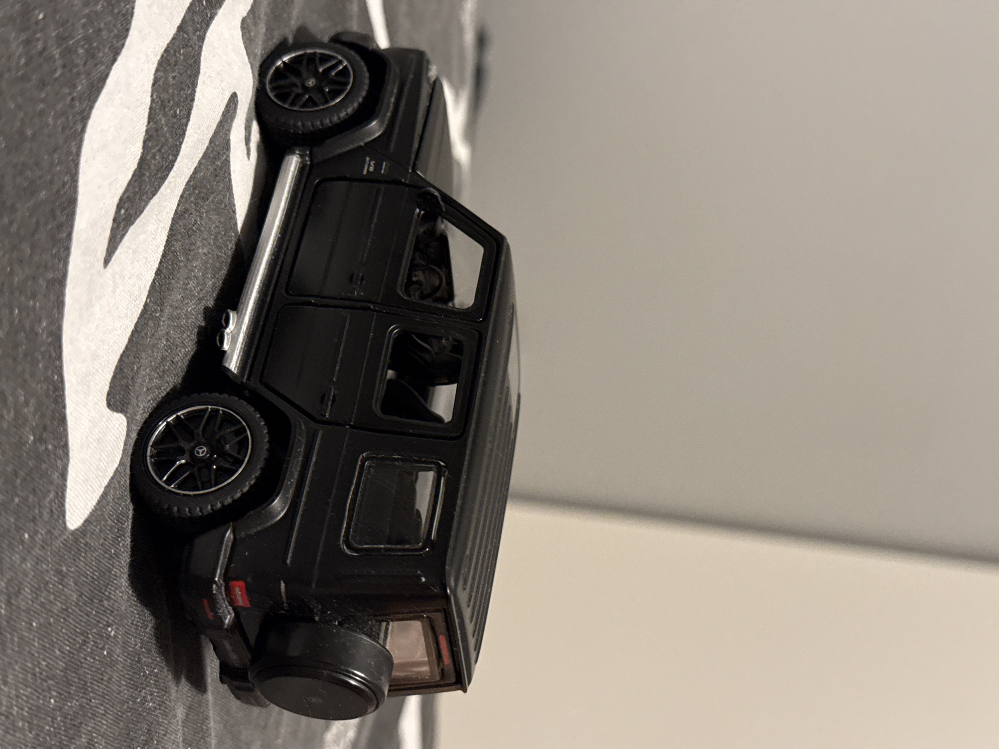 The image shows a black toy Mercedes-Benz G-Class SUV on a patterned surface. The toy car is detailed, featuring realistic wheels with the Mercedes-Benz logo, side mirrors, and a spare tire on the back. The surface it rests on appears to be a fabric with a bold, wavy, black and white stripe pattern. The background is a plain, light-colored wall, suggesting an indoor setting. The lighting is artificial, casting soft shadows. There is no visible text within the image. The scene is static, with the toy car simply positioned on the surface. The overall impression is that of a still life focused on a toy vehicle.