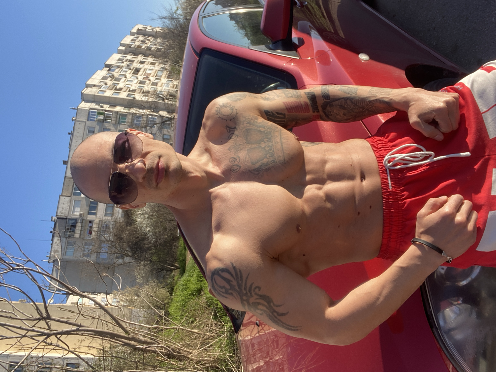 A shirtless bald man with sunglasses is posing next to a red car on a sunny day. He has numerous tattoos on his arms and chest. He is wearing red and white shorts and a black bracelet. In the background, there is a large apartment building complex, and some bare trees and grass, suggesting it may be early spring. The location appears to be in Pleven, Bulgaria, based on the typical architecture seen in the apartment building.