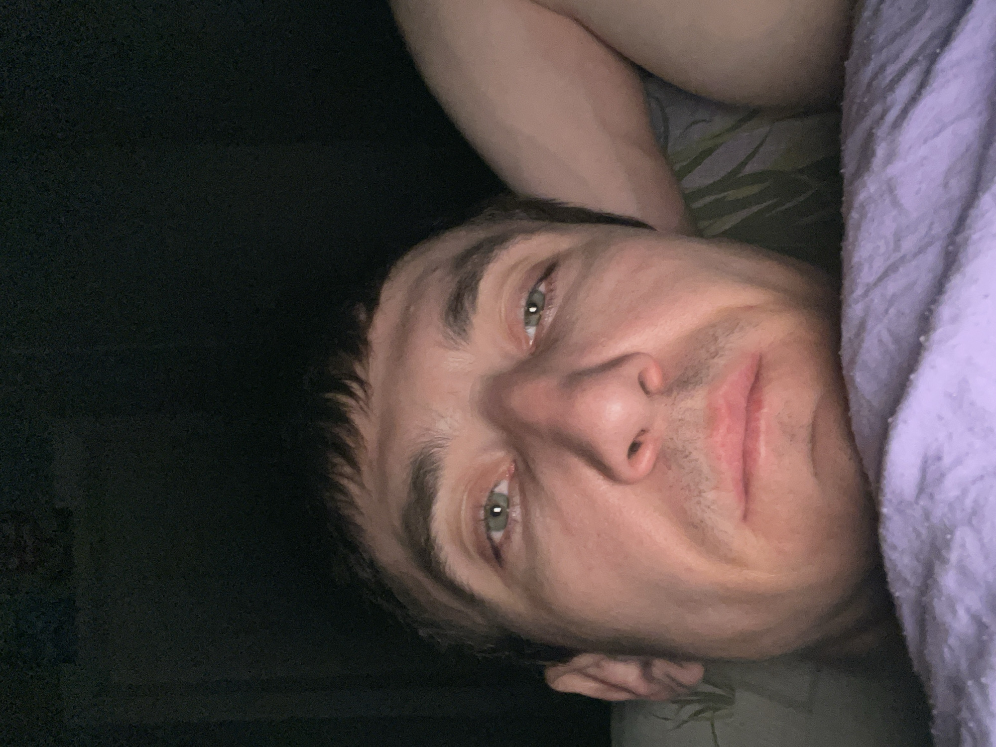 A close-up, low-angle, selfie-style photograph captures the face of a man lying down in what appears to be a bedroom. The man's head is positioned towards the top of the frame, with his gaze directed at the camera. His eyes are open and appear to be a light green color. He has short, dark hair with bangs. His skin tone is fair, and he has a few days' stubble on his chin and upper lip. His lips are slightly parted.
The man is lying on a bed. A portion of his right arm is visible, resting behind his head. The bedding appears to be a light purple blanket or comforter in the foreground, and a patterned sheet or pillowcase with green leaf designs is visible further back. The background is dark and out of focus, suggesting a dimly lit environment, possibly at night or with the lights off. There is no indication of the specific location within Laishevo, Russia, as the image does not contain any identifiable landmarks or details that would confirm its setting.