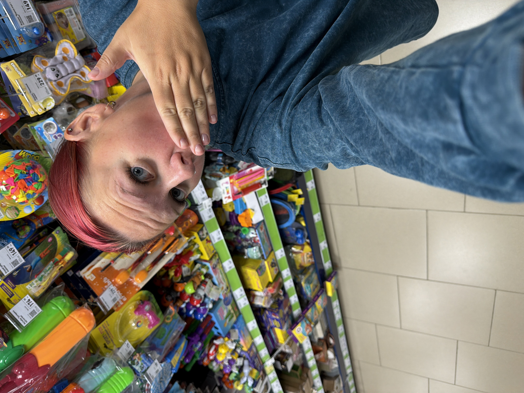 The image shows a young woman with pink hair and blue eyes, wearing a blue t-shirt. She is standing in a store aisle filled with shelves of toys. Her right hand is covering her mouth. The shelves are stocked with various colorful toys, including water guns, plush toys, and other novelty items. The floor is tiled with light beige square tiles. The overall lighting suggests an indoor retail environment. The scene appears to be in a toy store.