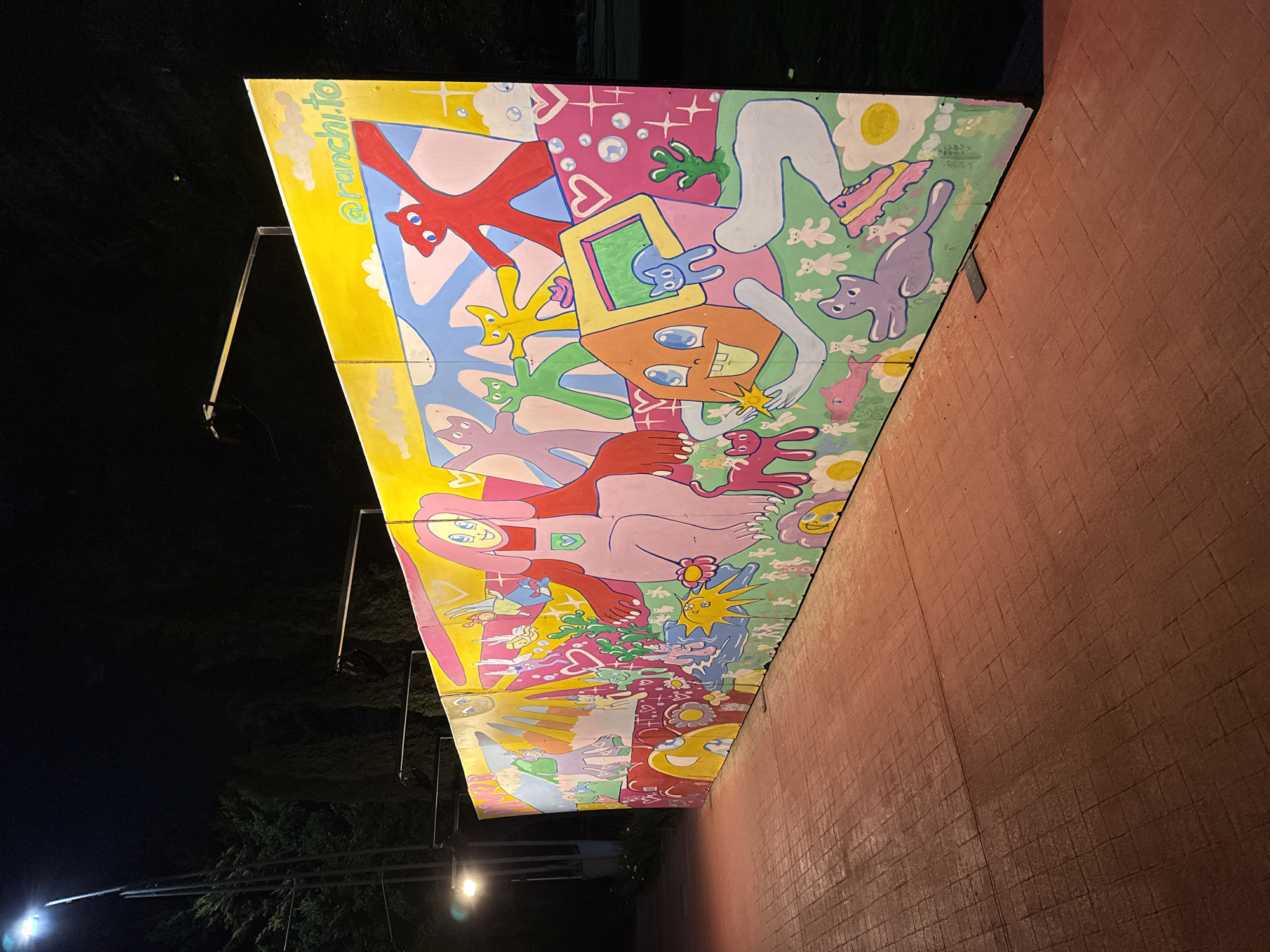 Here is a description of the image:
The image shows an outdoor mural located along a brick wall. The mural is brightly colored with cartoonish figures and shapes. The mural is supported by metal bars and is illuminated by streetlights. The location of the mural may be in Puebla, Mexico, as the mural shows "@ranchi.to."