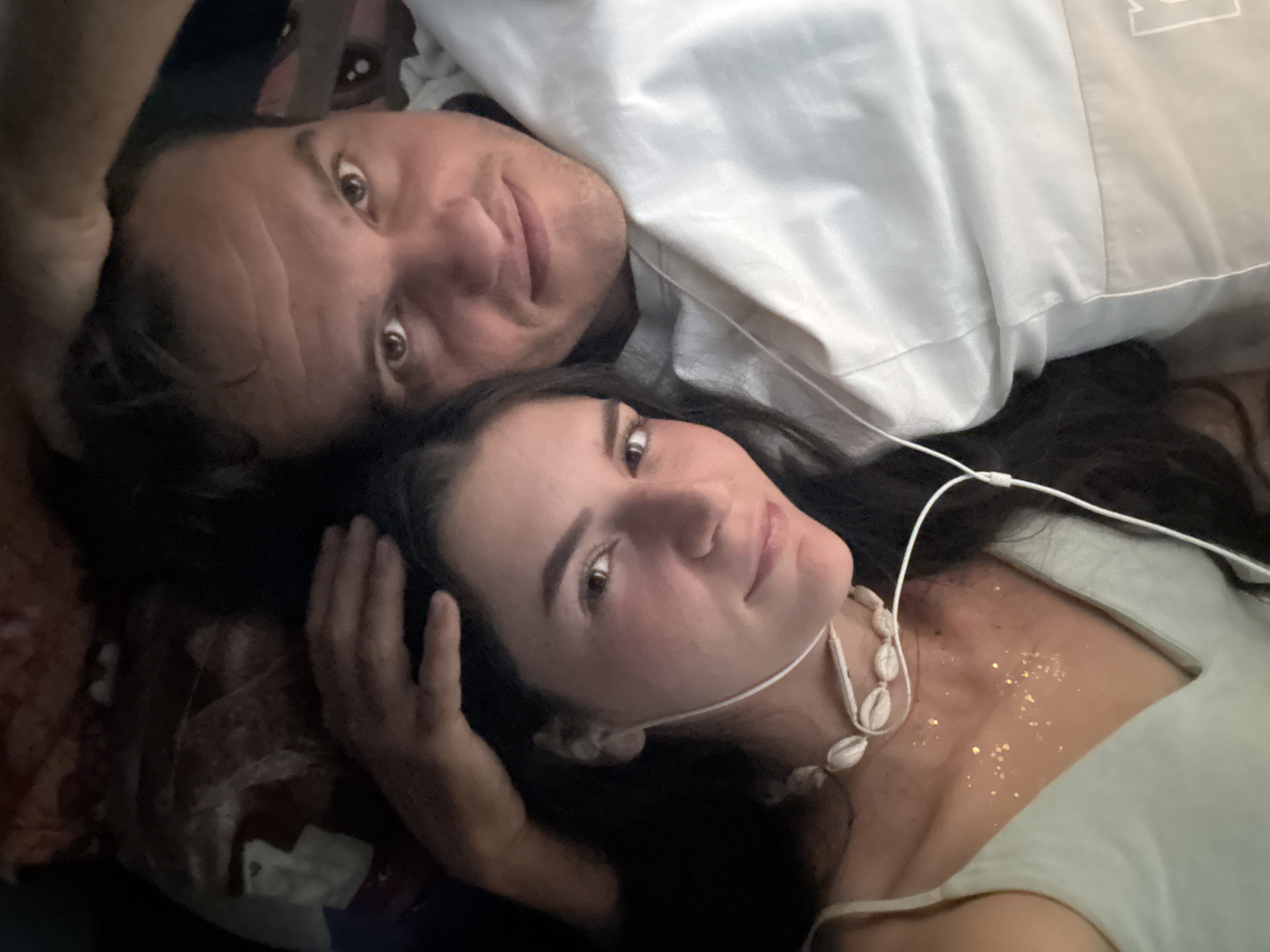 The image shows a man and a woman lying next to each other, looking up at the camera. The man has short, dark hair and is wearing a white shirt. The woman has long, dark hair and is wearing a light-colored top and a seashell necklace. Gold glitter is visible on her chest. A white wire, likely from headphones, is draped around her neck. The background is dark and indistinct. The location is Phu Thuy Ward, Vietnam.