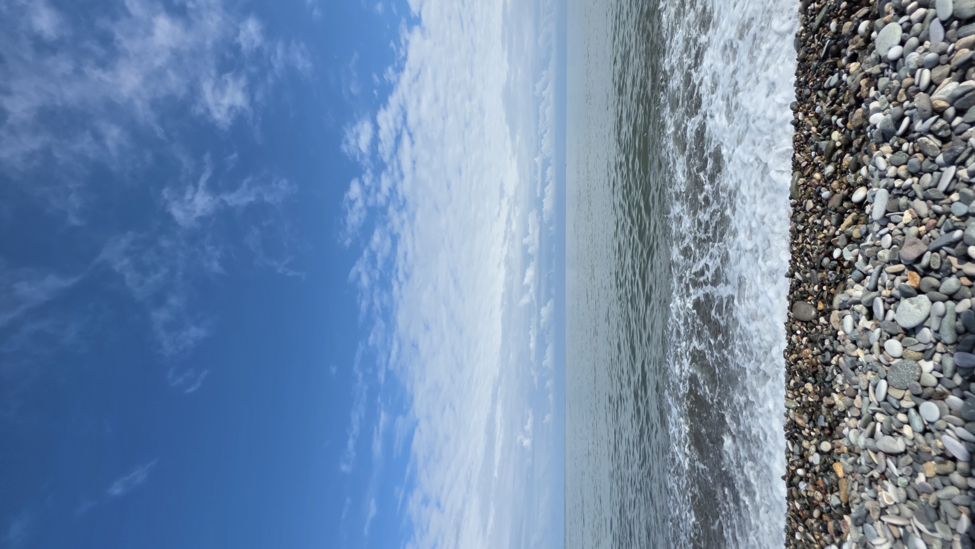 The image depicts a tranquil scene of the Black Sea coast. In the foreground, a dense bed of colorful pebbles covers the beach, with a gentle wave lapping at the shore, creating a frothy white edge. The sea itself appears calm, with a muted grey-green hue, stretching towards the horizon. Above, a vast expanse of blue sky is dotted with scattered, wispy white clouds, suggesting a pleasant day. The horizon line where the sea meets the sky is clear and defined. There are no people or man-made structures visible in the frame, creating an untouched, natural landscape. This scene is characteristic of many beaches in Batumi, Georgia, which is known for its pebble beaches along the Black Sea.