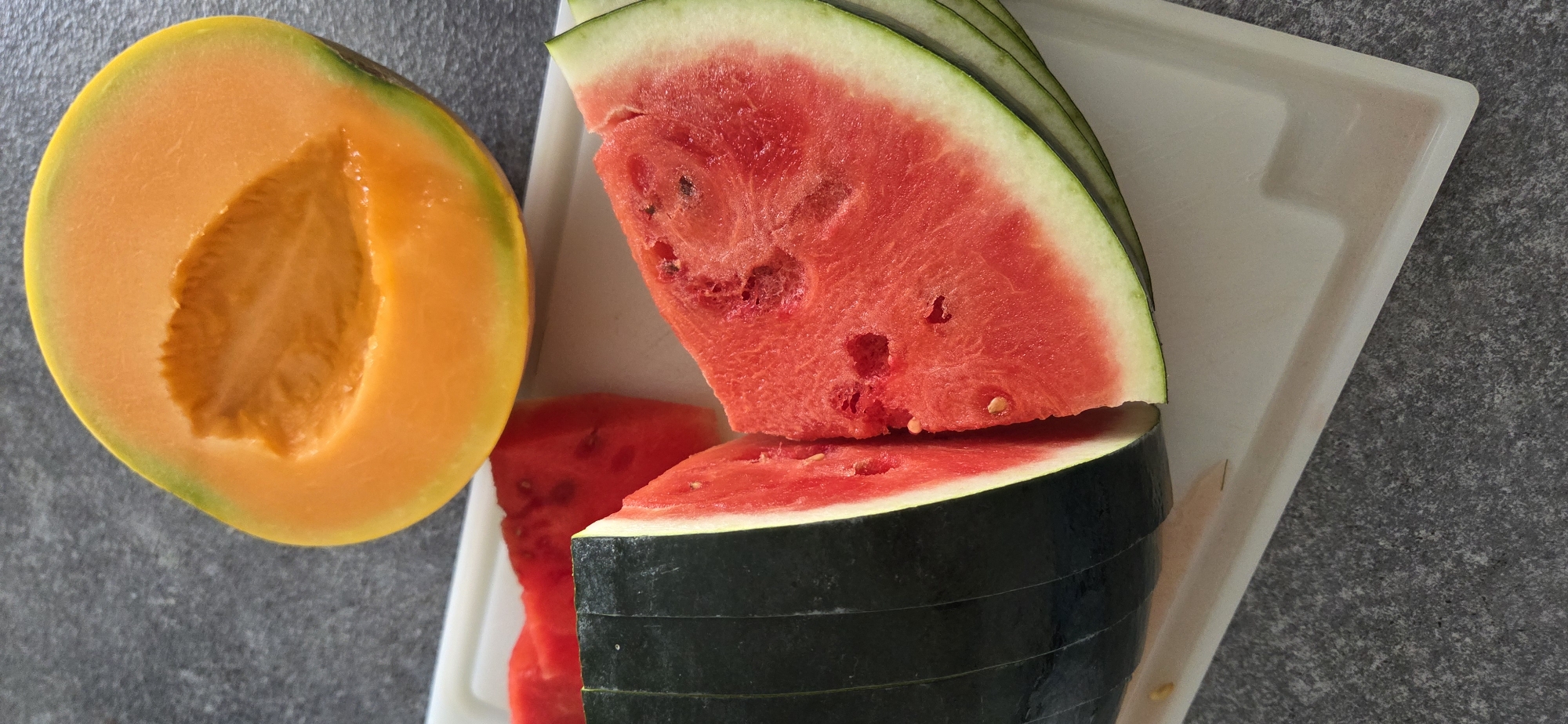 An overhead shot shows a whole cantaloupe and sliced watermelon on a white cutting board, which sits on a dark gray, speckled surface. The cantaloupe is a bright orange with a green rind, and it is cut in half, revealing the seedy interior. The watermelon is sliced into several wedges, with a dark green rind and bright red flesh. Some of the watermelon slices have been cut further into smaller pieces. The scene is brightly lit, with natural light casting soft shadows.