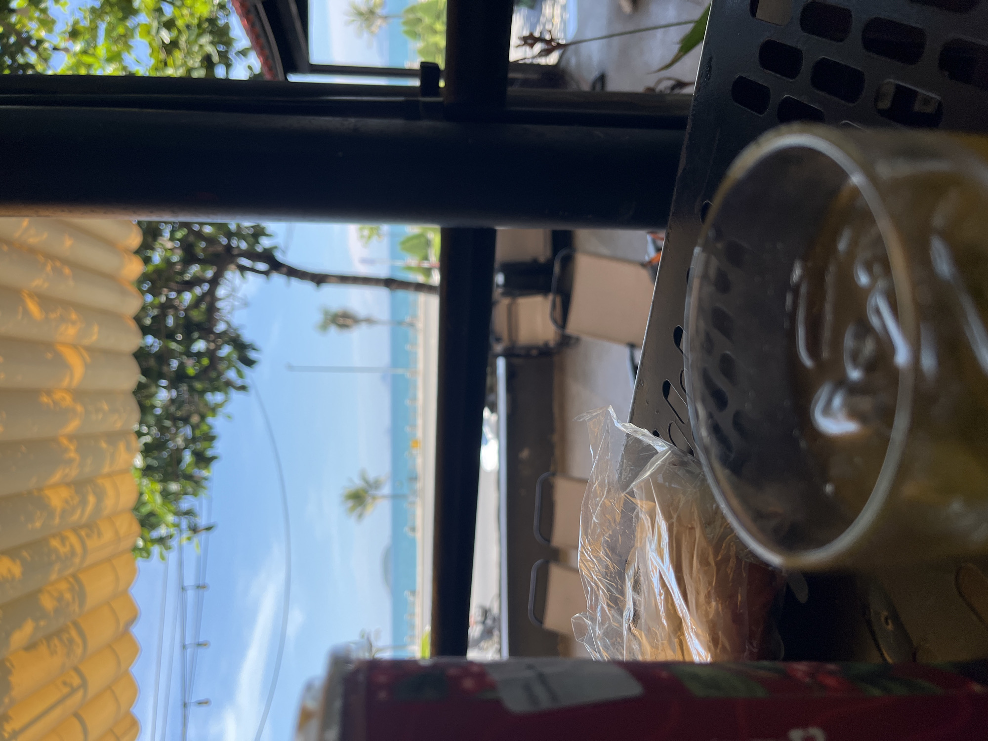 The image depicts a view from a shaded outdoor seating area overlooking a beach in Bac Nha Trang Ward, Vietnam. In the foreground, a glass of amber liquid with ice is visible, resting on a black metal grate. To the left, a portion of a red cylindrical object, possibly a can, is in focus. Behind the glass, a plastic-wrapped item, likely food, lies on the table. The background reveals a street with parked motorcycles, followed by a row of empty lounge chairs. Beyond the chairs, a palm-lined promenade separates the seating area from the ocean, which stretches to the horizon under a clear blue sky. A light yellow awning with shadow patterns covers the seating area.