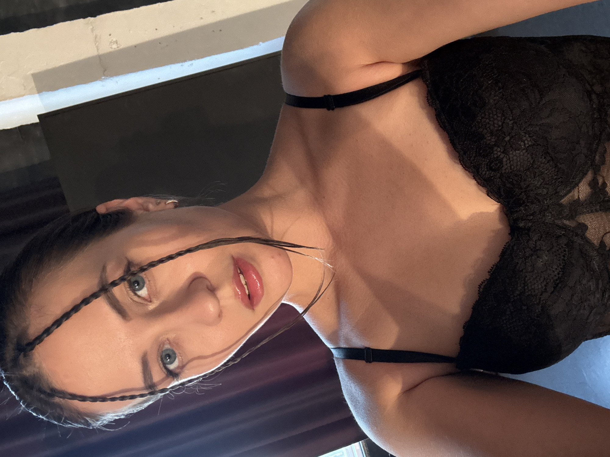 A young woman is photographed from a low angle, wearing a black lace bra. Her hair is styled with thin braids framing her face. The background is partially obscured, featuring elements of a room with a dark panel and a white and gray striped section. The location is not identifiable as Izhevsk, Russia based on the provided image.