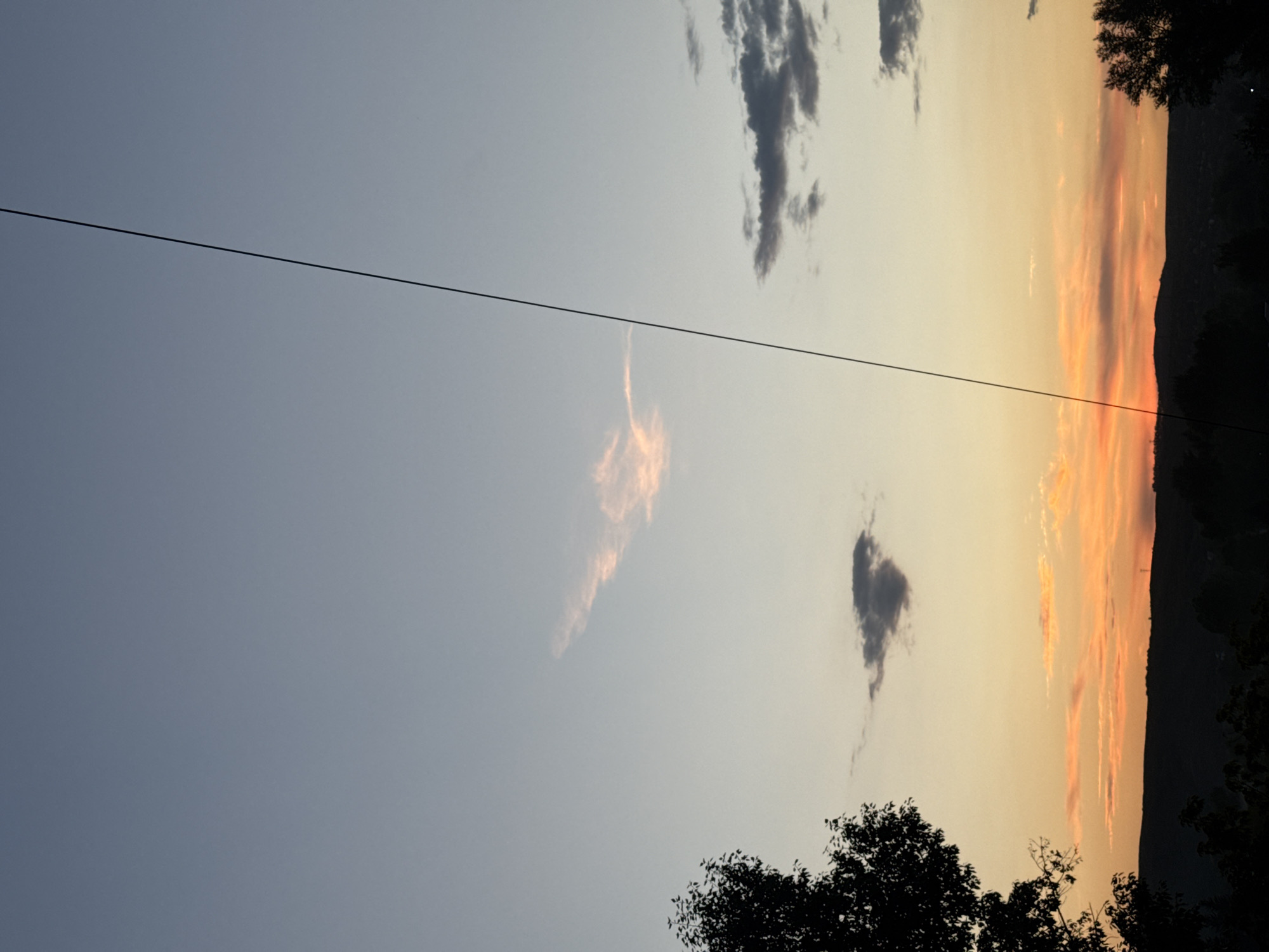 A silhouetted tree is visible on the left side of the frame, with its branches dark against the soft gradient of the sky. A thin, dark power line bisects the image diagonally from the top right towards the bottom. The sky displays a gentle transition from a pale blue at the top to hues of orange and yellow near the horizon. A few wispy clouds are scattered across the sky; one in the upper center is tinged with pink, and another darker, heart-shaped cloud is located below it. In the distance, a dark, undulating silhouette of hills or mountains stretches across the horizon, dotted with more trees and some faint lights, suggesting a rural or suburban landscape. The overall scene captures the serene beauty of a sunset.