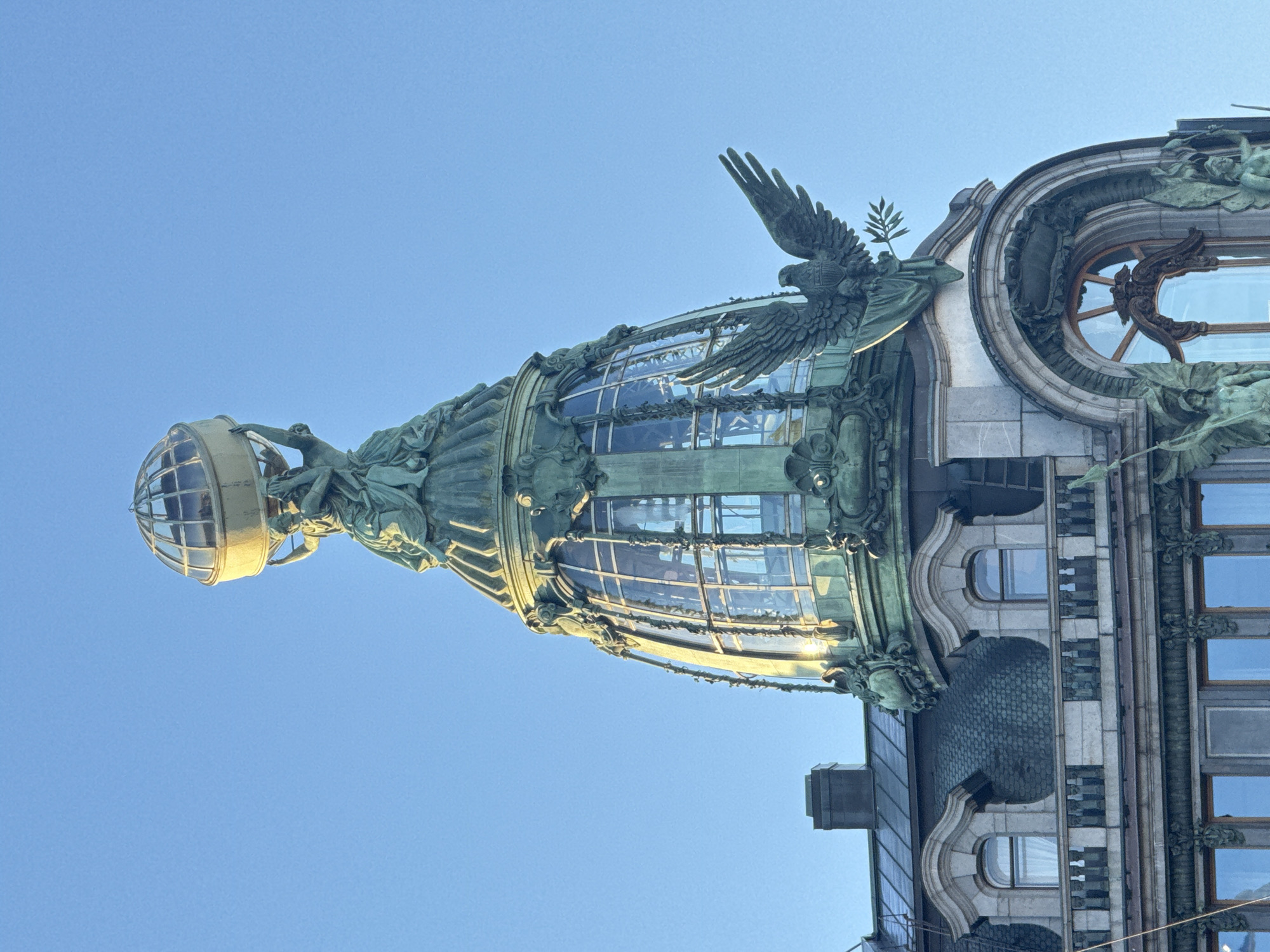 The image shows a portion of the Singer House, a landmark building located at Nevsky Prospect 28 in Saint Petersburg, Russia. The photograph focuses on the architectural details of the building, particularly the corner tower topped with a glass dome. The dome is adorned with sculptures of figures, including a woman holding a globe, and an eagle perched on the side with a branch in its beak. The lower part of the image includes a section of the building's facade, which features windows, stone cladding, and more sculptural elements. The background consists of a clear blue sky.