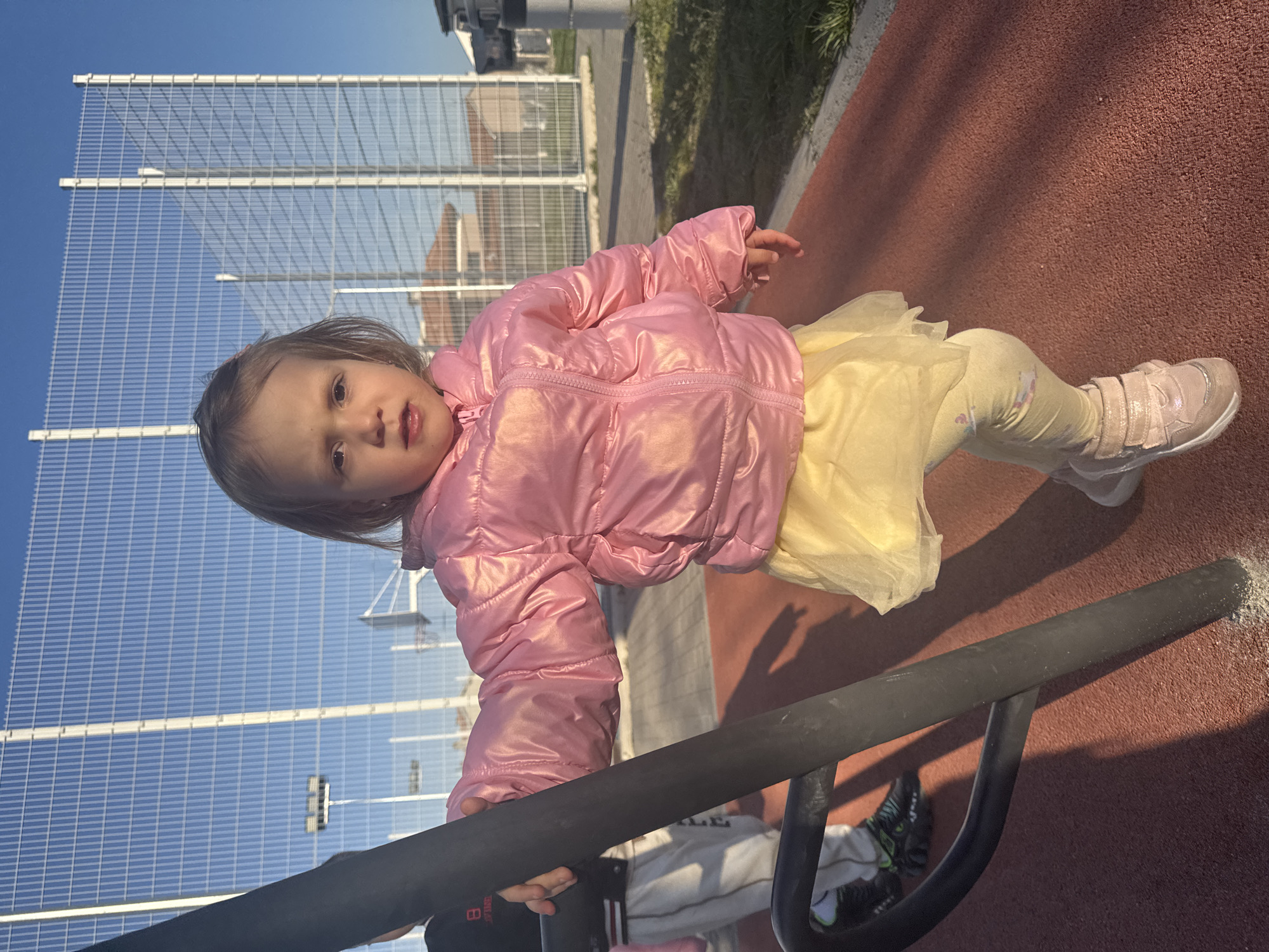 A young child, likely between 2 and 4 years old, is holding onto a piece of black playground equipment, possibly monkey bars. She is wearing a shiny pink jacket, a yellow tutu-like skirt, patterned leggings, and light-colored sneakers. The child is on a red, textured surface typical of playgrounds. In the background, there is a tall, wire mesh fence and buildings. The sky is bright blue. The scene is likely located in Varna, Bulgaria, although there are no specific indicators in the image to confirm.