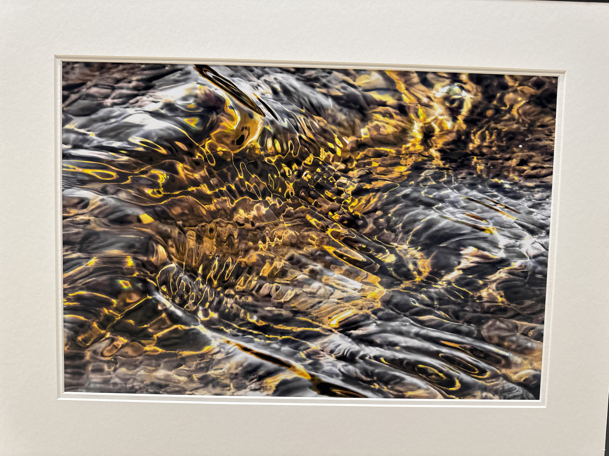 This is a close-up, abstract photograph of flowing water, framed by a white mat. The water exhibits a dynamic play of light and shadow, with shimmering golden hues reflecting from an unseen light source against a backdrop of dark, rippling textures. The surface of the water is highly textured, with waves and eddies creating intricate patterns. The scene appears to be a natural water feature, possibly a stream or river, with submerged stones or substrate visible beneath the disturbed surface. The context of Kilkenny, Ireland is not evident within the image itself. There are no people, animals, or man-made structures visible.
