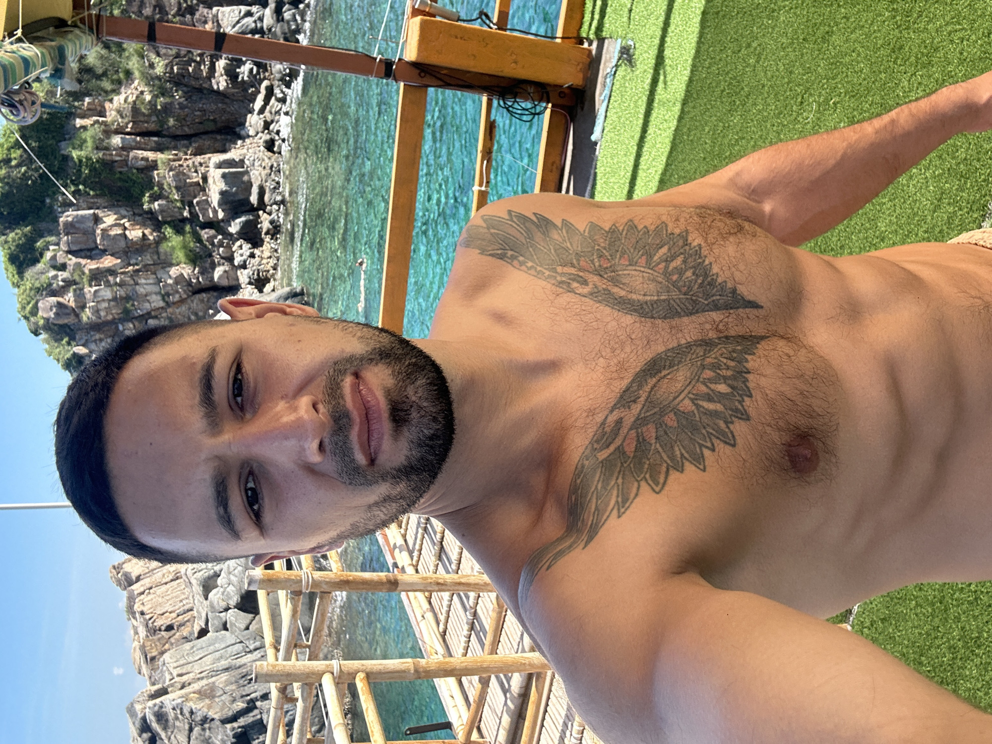 A man with dark hair and a beard takes a selfie on a boat. He is shirtless and has a tattoo of wings on his chest. He is standing on a deck covered in green turf, with a wooden railing and the ocean in the background. The ocean is a vibrant turquoise color, and rocky islands with lush greenery can be seen in the distance. The sky is clear and blue. The scene appears to be in a tropical location. The man is looking directly at the camera with a neutral expression.