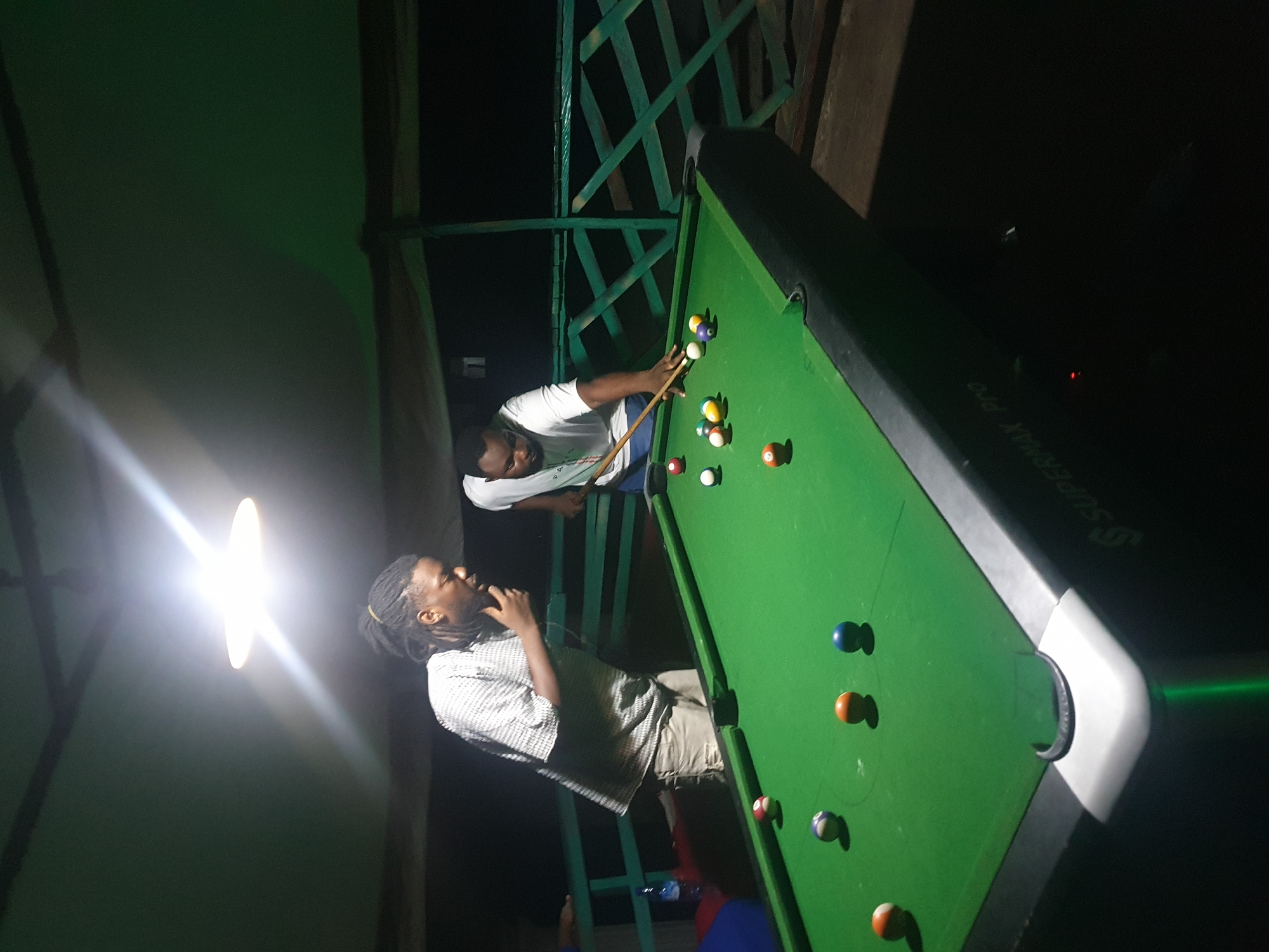 Two men are playing pool at a rooftop bar in Lokoja, Nigeria. The man on the left is wearing a short-sleeved, checkered shirt and light-colored shorts, with his dreadlocks pulled back in a bun. He is observing the game with his hand on his chin. The man on the right, wearing a white t-shirt and blue shorts, is in the process of striking a billiard ball with a cue. The billiard table is green and has several balls scattered on it. In the background, there is a railing, suggesting the location is elevated. The scene is lit by a bright overhead light.