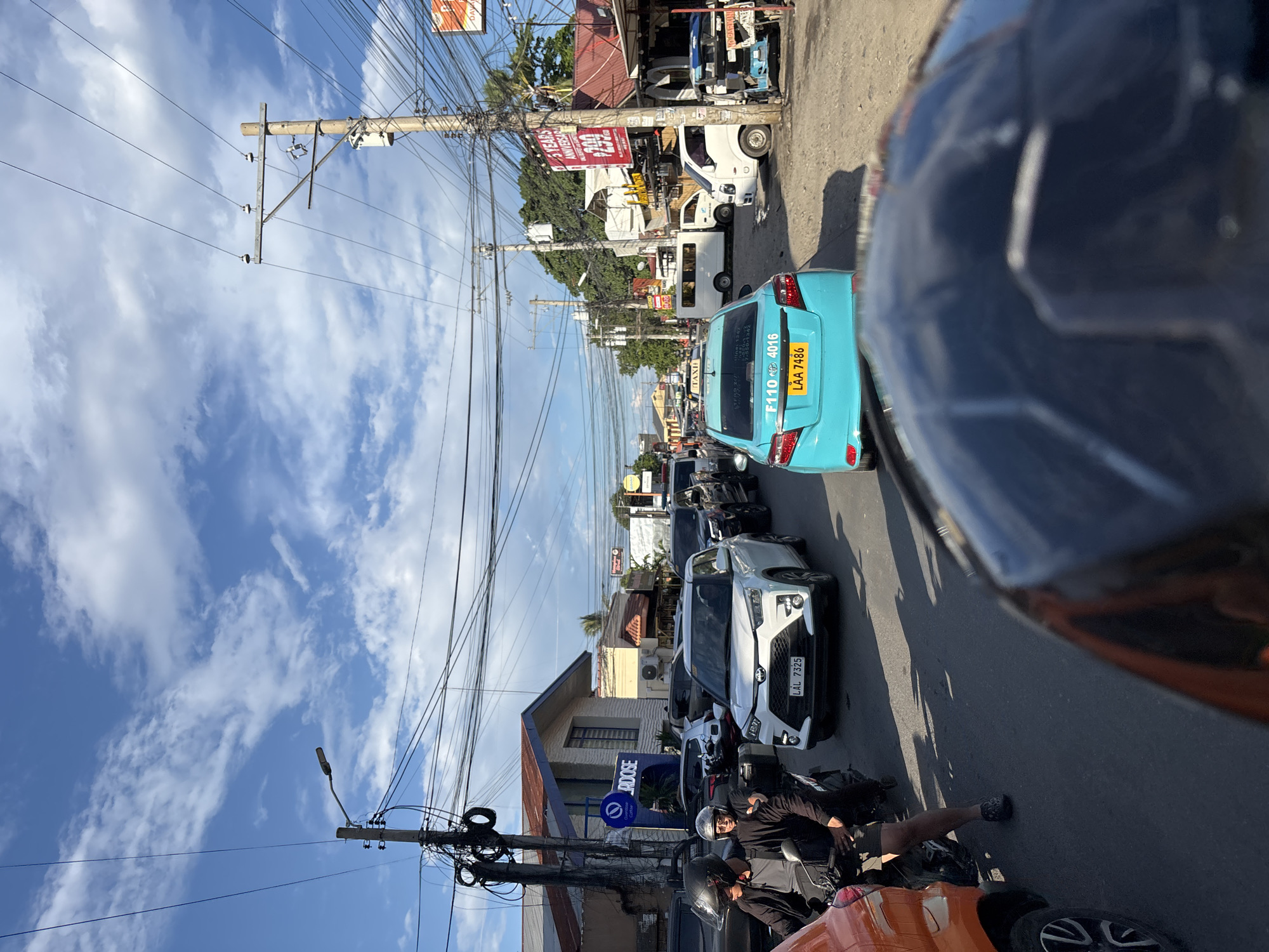 The image captures a traffic scene on a street in Davao City, Philippines, under a partly cloudy sky. In the foreground, the top of a dark helmet is visible, suggesting the photo is taken from the perspective of someone riding a motorcycle.
A white SUV, with license plate LAL 7325, is prominently positioned in the middle of the frame, stopped in traffic. To its left, two individuals on a motorcycle are also stopped, the driver wearing a black jacket and helmet, and the passenger wearing a silver helmet and dark clothing. The passenger appears to be looking forward.
Further down the street, a turquoise sedan, likely a taxi, with license plate LAA 7486 and markings "F110 4016", is also part of the congestion. Several other vehicles, including a white van and a white utility vehicle, are visible in the background, as well as a blue tricycle.
The environment includes buildings lining the street, some with signage such as "DOSE Coffee" and another displaying a "5 YEARS ANNIVERSARY" banner with a price of "299". Utility poles with a dense network of electrical wires crisscross the sky, typical of urban Philippines. The road appears to be paved with asphalt, and shadows from the vehicles and poles are cast on the ground, indicating it is daytime. The overall impression is one of typical urban traffic in the Philippines, likely during a busy period.