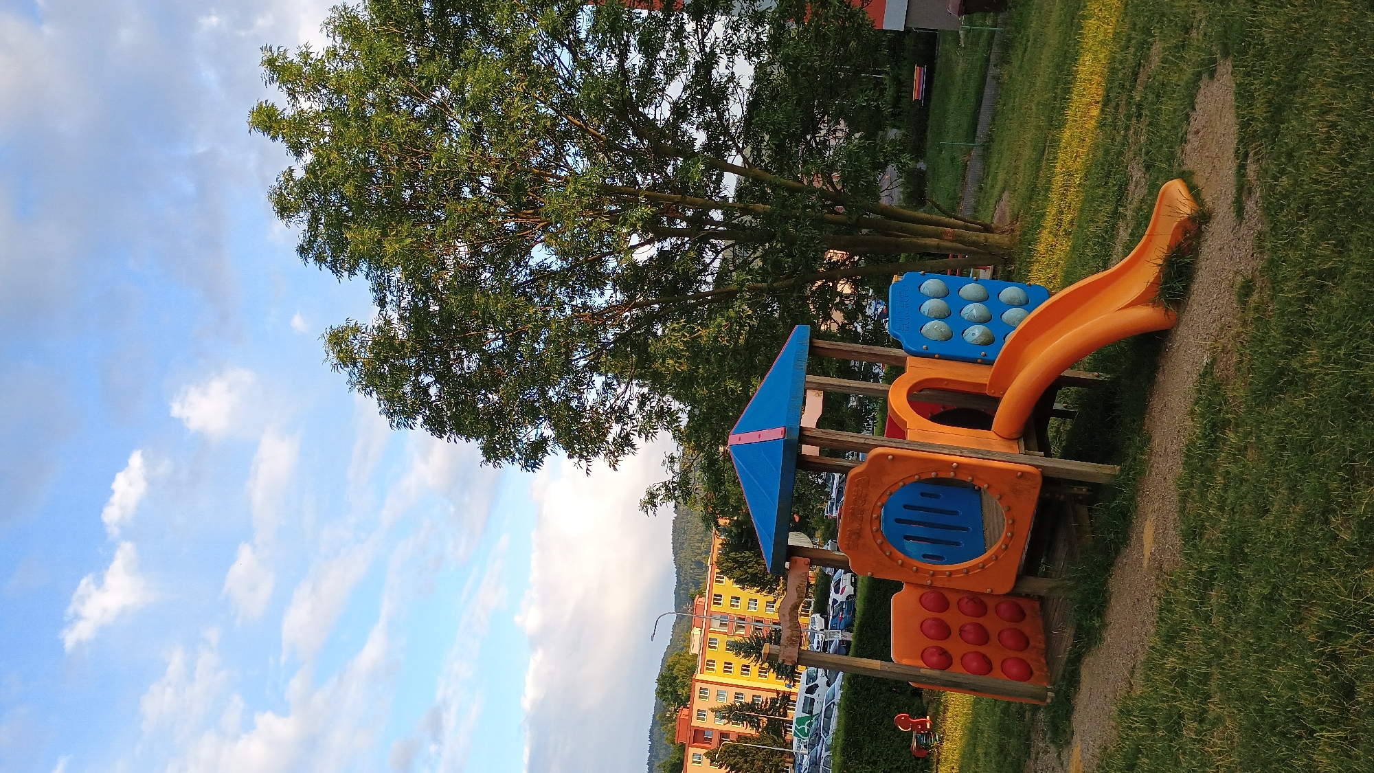 The image shows a playground structure with a blue and pink roof, orange and blue panels, and an orange slide. The playground is situated on a grassy area with a dirt path leading to it. In the background, there are apartment buildings with yellow and orange facades, parked cars, and a large tree. The sky is blue with scattered white clouds. The scene appears to be in a residential area of Sokolov, Czechia, during daytime. There are no people visible in the image.