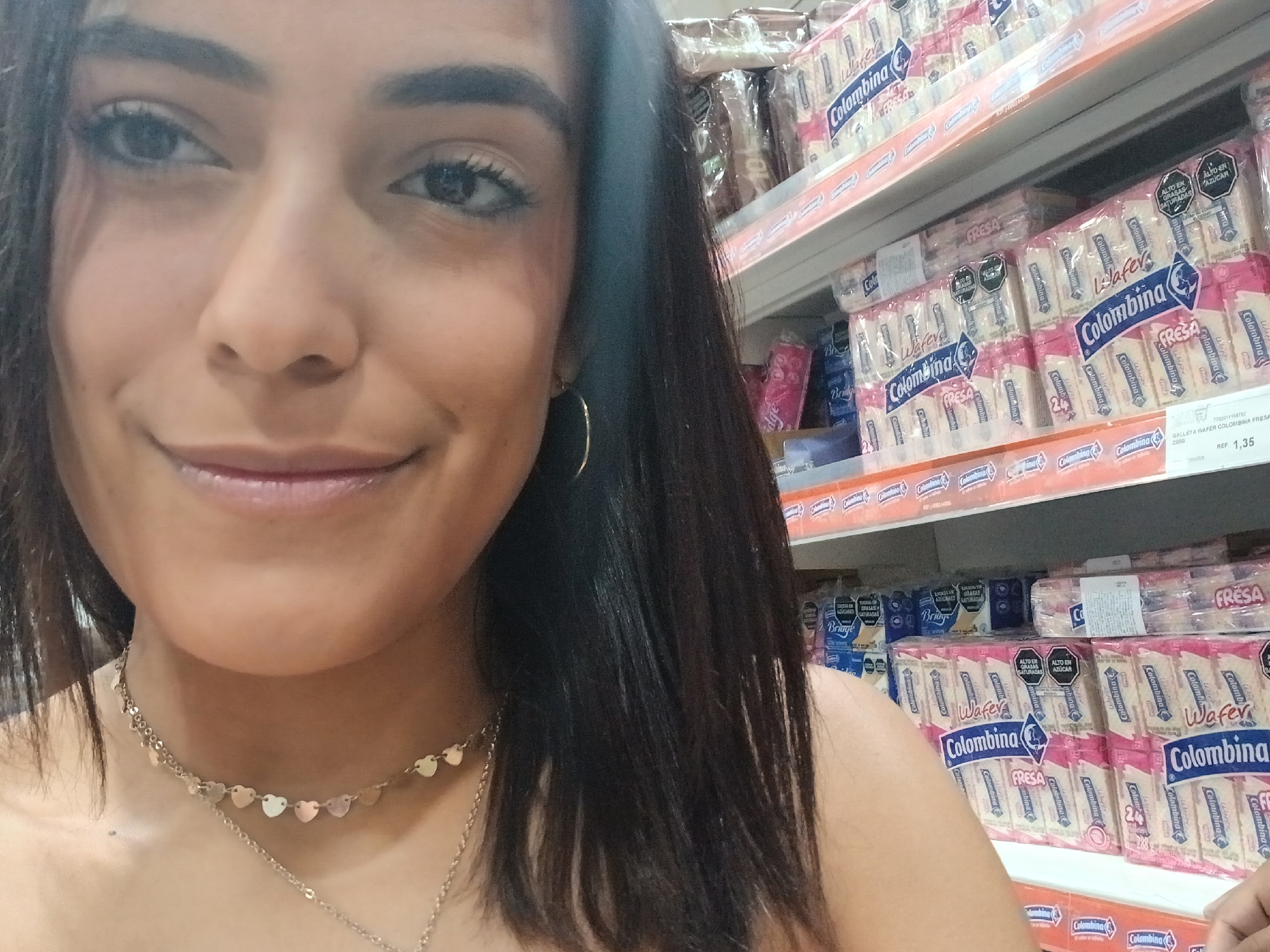 A woman with dark hair and a heart-shaped necklace is taking a selfie in a supermarket. The shelves behind her are stocked with packages of Colombina wafer cookies, labeled "Fresa" (strawberry flavor). The cookies have black warning labels indicating high levels of saturated fats and sugar. A price tag for 1.35 currency is visible. The image provides no information that identifies the location as Juan Griego, Venezuela.