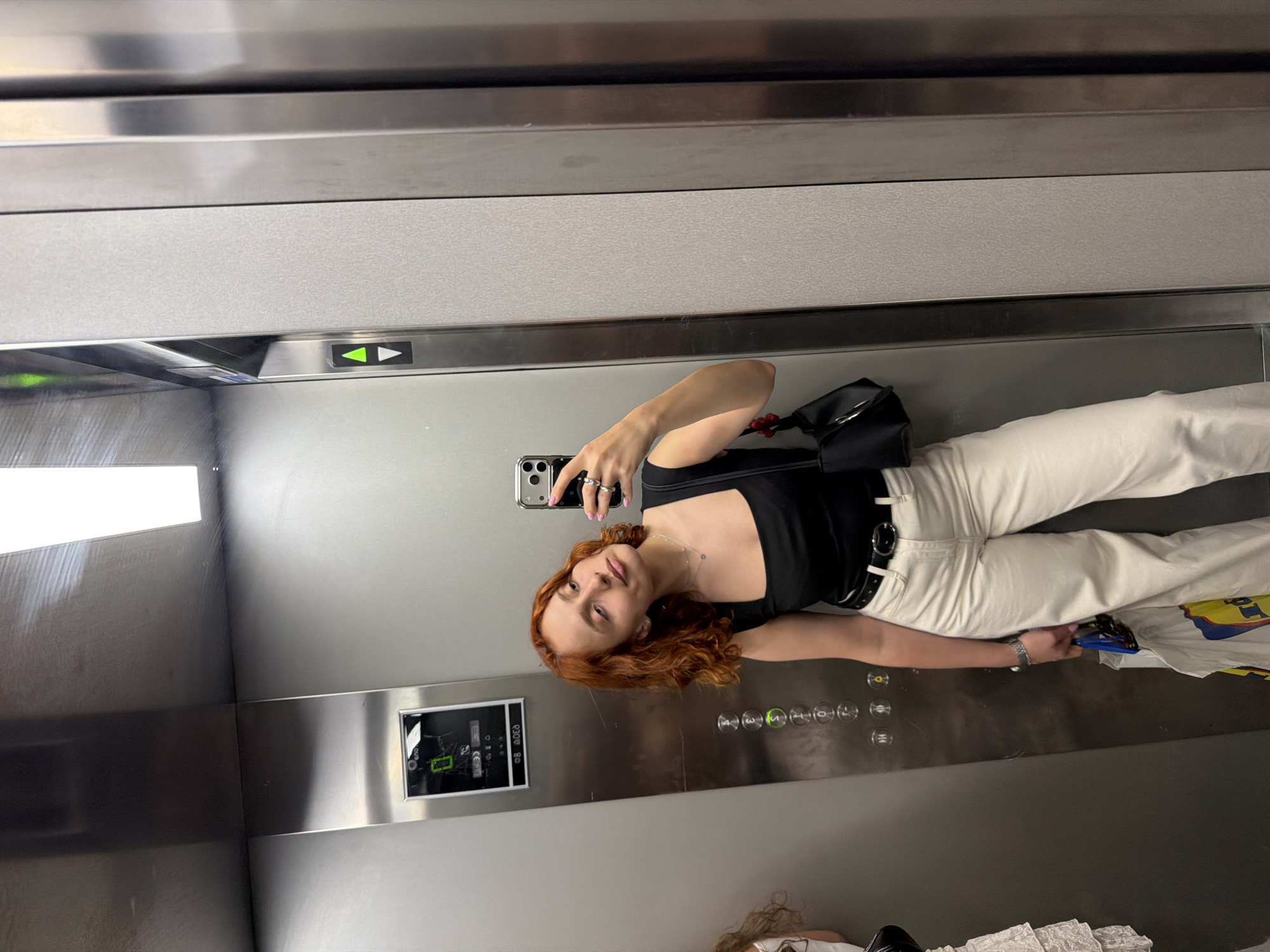 A young woman with reddish-brown hair, wearing a black tank top and light-colored pants, takes a mirror selfie in an elevator. She is holding a smartphone in her right hand, and a black purse hangs from her shoulder. A yellow shopping bag is partially visible in her left hand. The elevator has silver walls and a control panel with buttons. An illuminated green arrow is visible on the elevator wall. The scene takes place inside an elevator, likely in a building in Strasbourg, France.