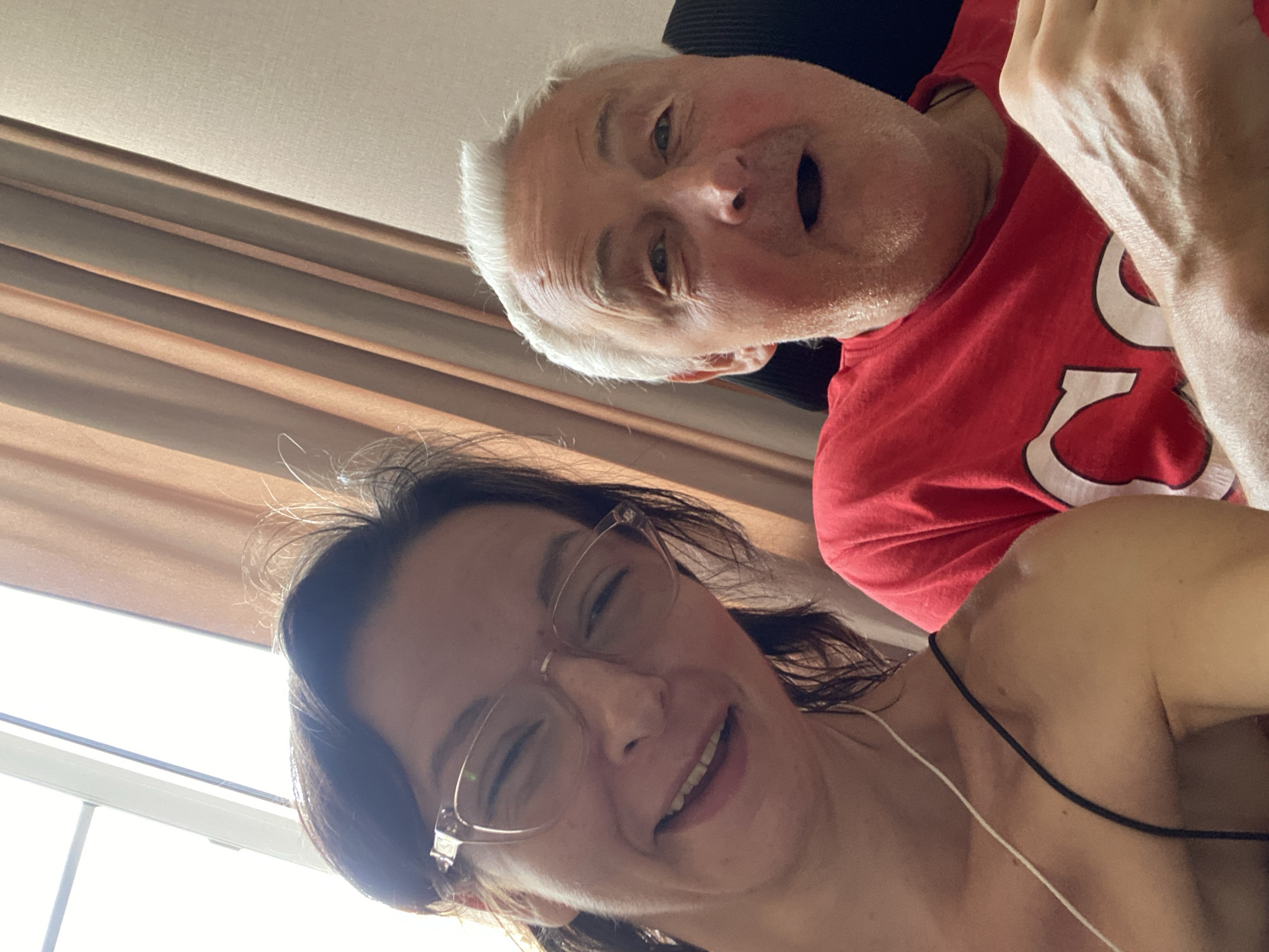 In a selfie taken indoors, a woman with dark hair and glasses smiles widely, her eyes crinkled. To her right, an older man with white hair and a red t-shirt looks at the camera with his mouth slightly open. The background consists of beige curtains and a window letting in bright light. The setting is likely a residential interior, with no specific markers indicating Yuzhny, Russia.