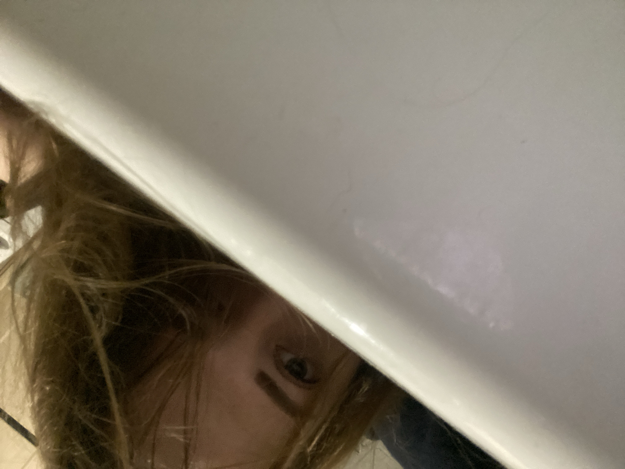 The image shows a close-up, low-angle shot of a person's face, peeking out from under a white surface. The person has light brown hair that is spread out around their face. Their left eye is visible and looking directly at the camera. There is a dark streak or mark on their cheekbone below the eye. The white surface appears to be a large, smooth object, possibly a table, counter, or lid, casting a shadow over much of the person's face. The lighting is dim, suggesting an indoor setting, possibly with artificial light. The overall mood is somewhat playful or secretive due to the way the person is positioned. There is no visible text in the image.