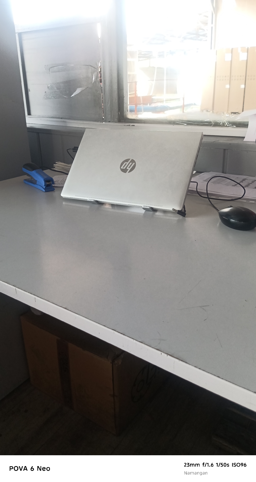 The image shows a desk with a laptop, a mouse, and a stapler. The laptop is a silver HP model. The mouse is black and wired. The stapler is blue. There are also some papers on the desk. In the background, there is a window with sunlight streaming through. Outside the window, there are some cardboard boxes. The image is taken in Namangan City, Uzbekistan. The lighting conditions suggest it is daytime. The scene appears to be an office or a workspace. There are no people visible in the image.