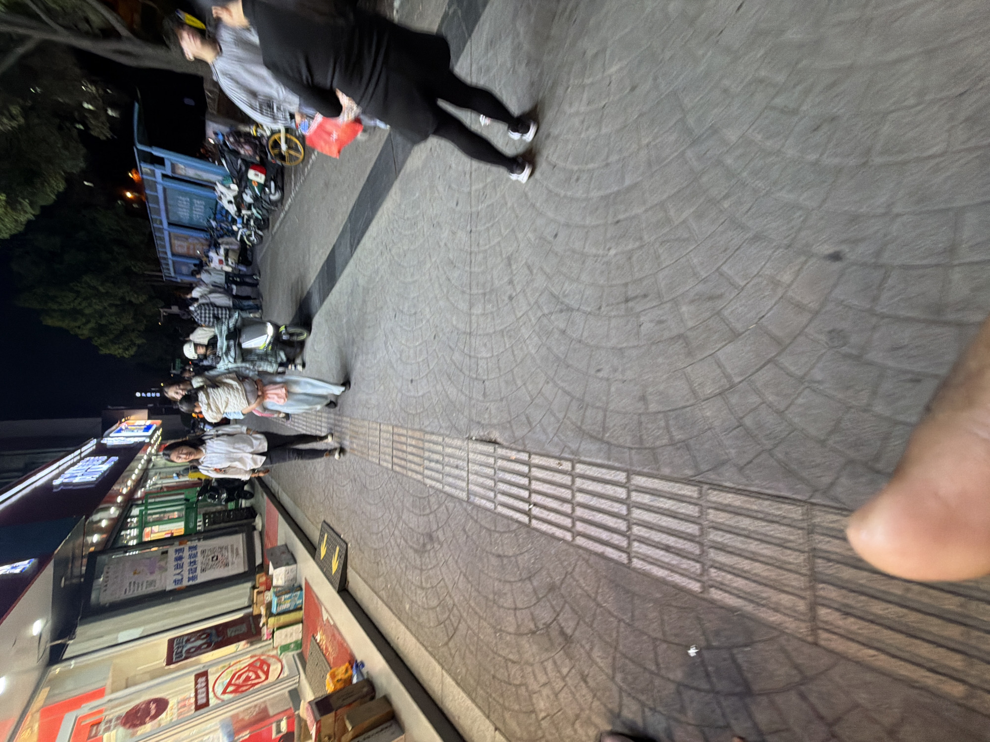 A street in Jiangning District, China, features shops on the left side of the frame, with pedestrians on the sidewalk. The street surface consists of rounded brick patterns and a textured guide path. A person is walking at the top of the frame, away from the camera. A line of parked bicycles is visible further back.