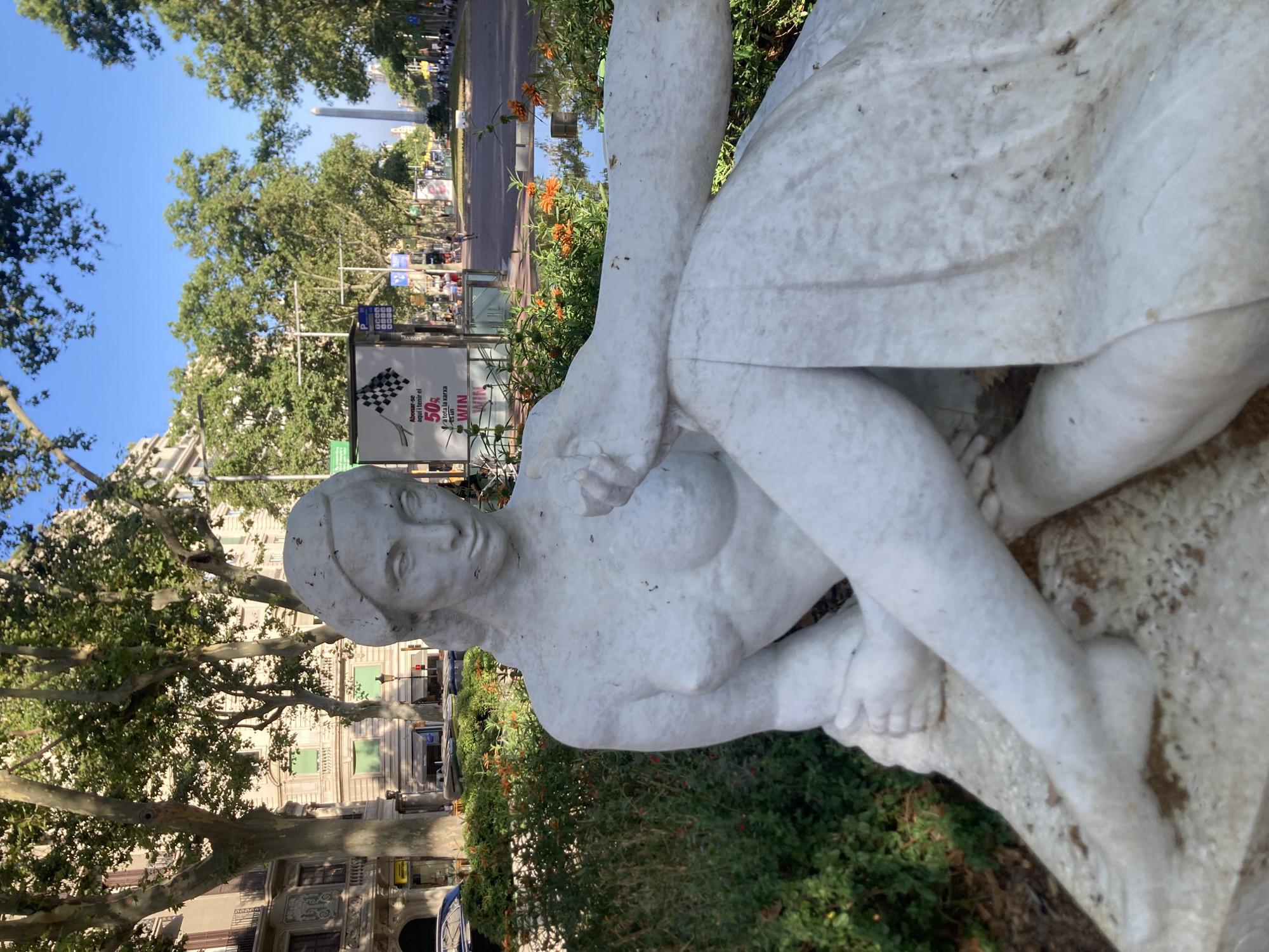 This image shows a white marble statue of a reclining woman in a public outdoor space. The statue is positioned in the foreground, with its head and upper body clearly visible, while the rest of its form is partially obscured by greenery and the edge of the frame. The woman in the statue has her legs crossed and her arms resting on her body, creating a relaxed and serene pose. Her facial features are delicate and serene.
In the background, a street scene unfolds with trees lining the sidewalk and buildings visible in the distance. There are indications of activity on the street, with a few figures walking and what appears to be a monument or tall structure in the far distance. A billboard is also visible, displaying a checkered flag and text in what appears to be Catalan, advertising a 50% discount. The lighting suggests it is a bright, sunny day. The overall atmosphere is one of tranquility, contrasting with the bustling urban environment suggested by the background. The presence of the statue and the billboard suggests a public park or a plaza in a city.