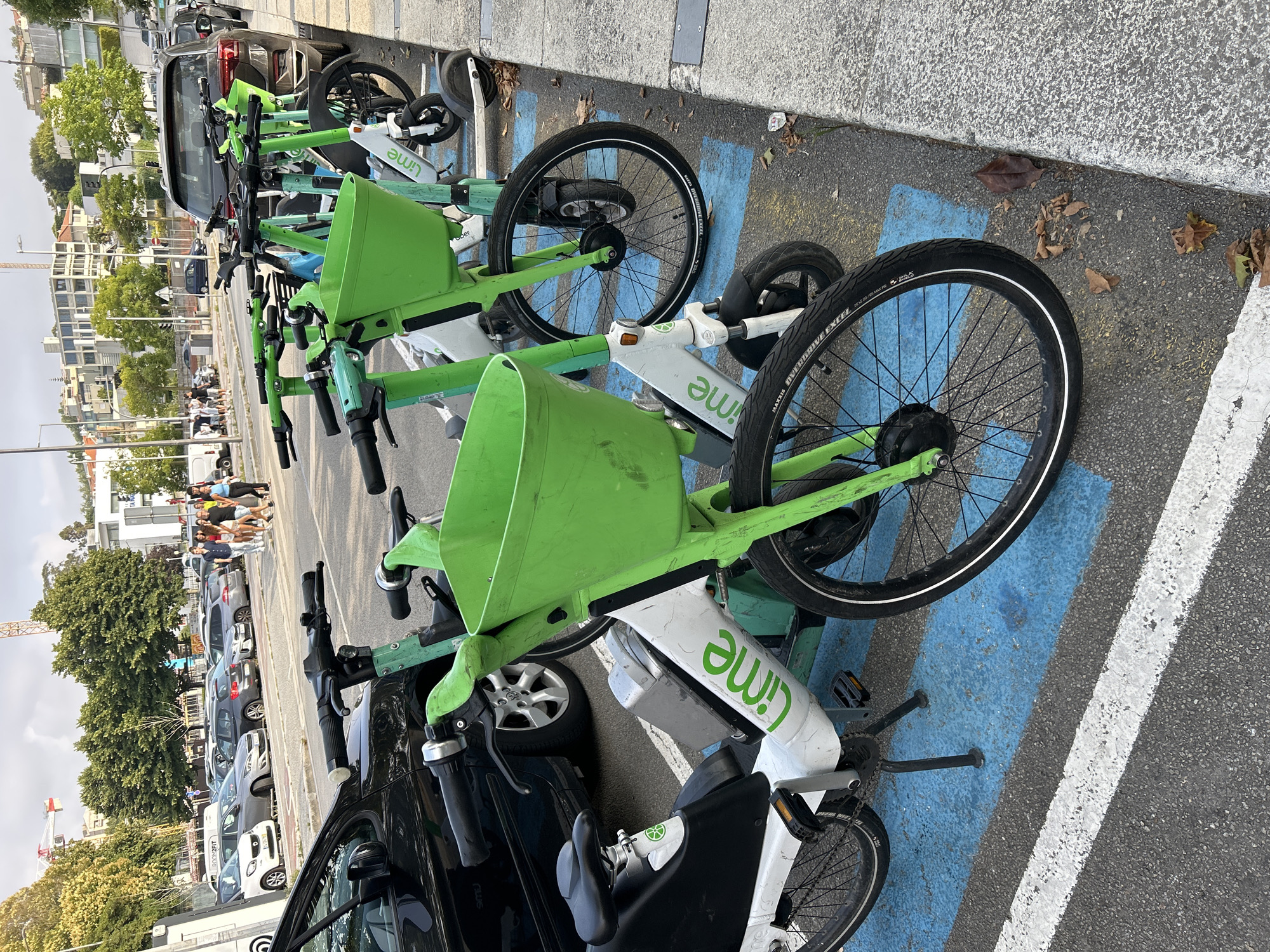 The image shows a row of rental Lime scooters parked on a street in Porto, Portugal. The scooters are primarily white and green, with large green baskets at the front. They are parked in marked parking spots, indicated by blue and white stripes on the asphalt. A black car is parked to the left of the scooters, and other vehicles and buildings are visible in the background. A few people are walking in the distance. The sky is overcast with some clouds.