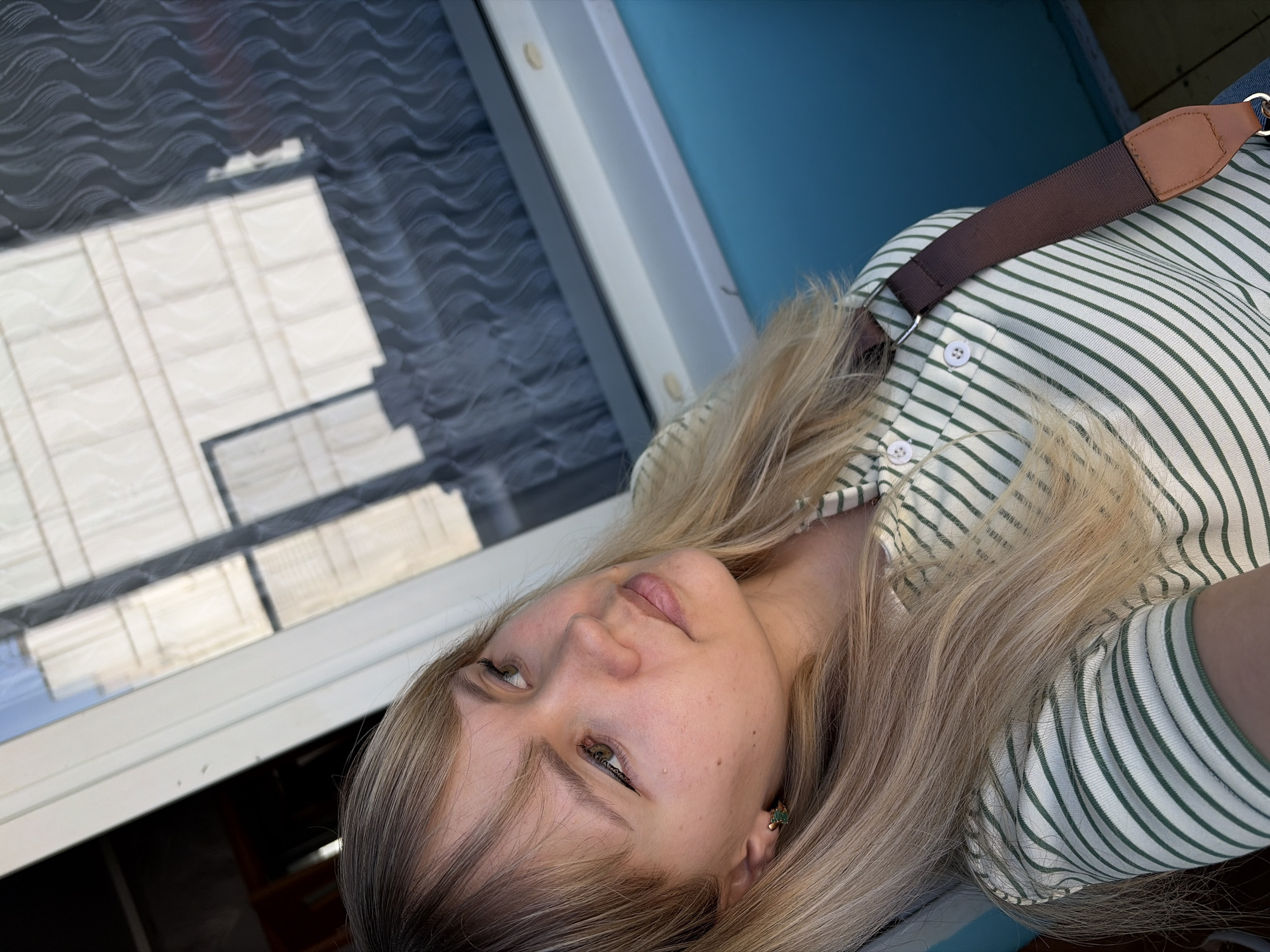 A light-skinned woman with blonde hair is angled in the foreground. She is wearing a striped shirt with a brown strap going across her shoulder. In the background, there is a window that reflects what appears to be a building or structures with a grid-like pattern. A blue wall or surface is visible to the right of the window frame. The precise location in the city of Chaikovsky, Russia, cannot be determined based solely on this image.
