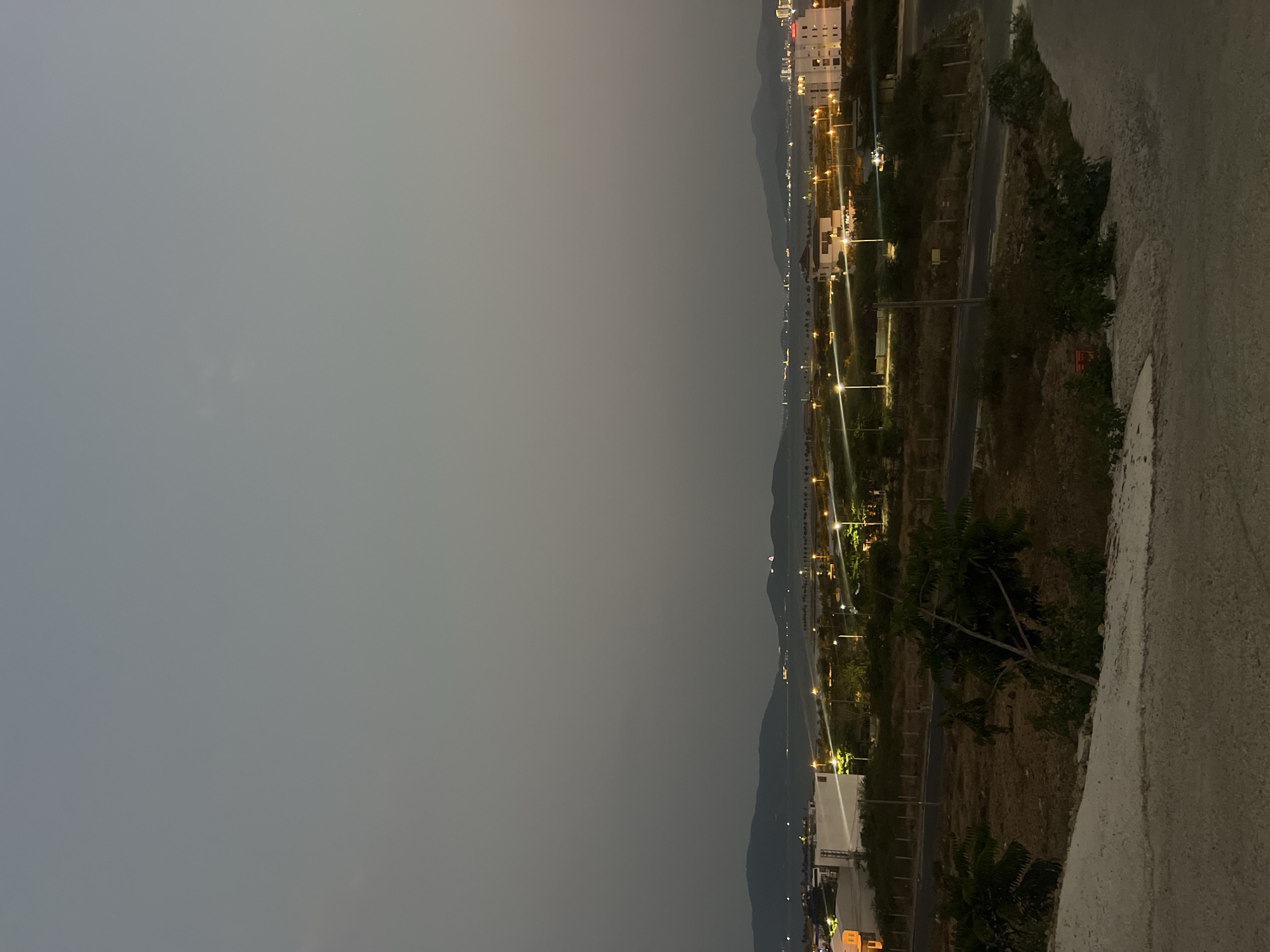 An elevated, wide-angle, night view of the city of Bắc Nha Trang, Vietnam, shows a portion of the city stretching along the coast. A mountain range serves as a backdrop along the coastline. A series of lights, presumably streetlights, run parallel to the coast. Buildings are visible, particularly in the far distance. In the foreground, a concrete barrier is seen with vegetation.
