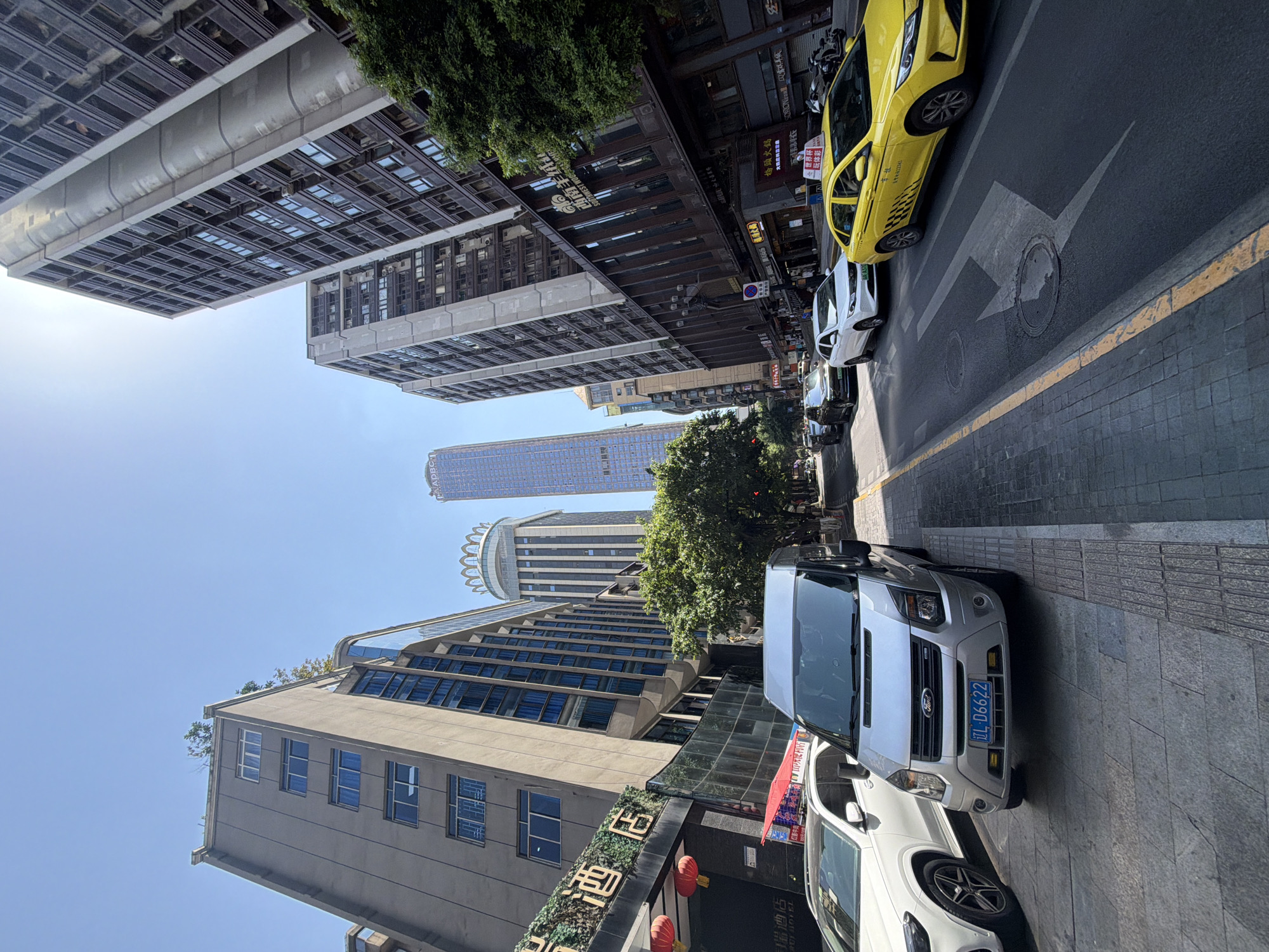 The image depicts a street scene in Yuzhong District, China, under a clear blue sky. The viewpoint is from a low angle, looking upwards towards tall buildings. On the left, a white SUV is parked on a paved sidewalk next to a silver Ford Transit van, also parked. The van's license plate reads "DL D6622". Further down the sidewalk, there is signage in Chinese characters, including "酒 庄", indicating a wine or liquor store, and red lanterns.
The street is occupied by several vehicles in motion. A yellow taxi is in the right lane, with Chinese text and a checkered pattern on its side. To its left, a white sedan and a dark-colored SUV are driving. The road has yellow lane markings and a large arrow-shaped marking pointing to the right on the asphalt, overlaying a manhole cover.
The buildings are modern and varied in design. On the left, a building with blue-tinted windows rises several stories. Behind it, another tall building with a distinctive crown-like structure at the top is visible. To the right, a series of very tall, narrow residential or office buildings with many windows are prominent, casting shadows on the street. Green trees line the right side of the street, obscuring some of the lower levels of the buildings. The overall impression is a bustling urban environment during daytime.